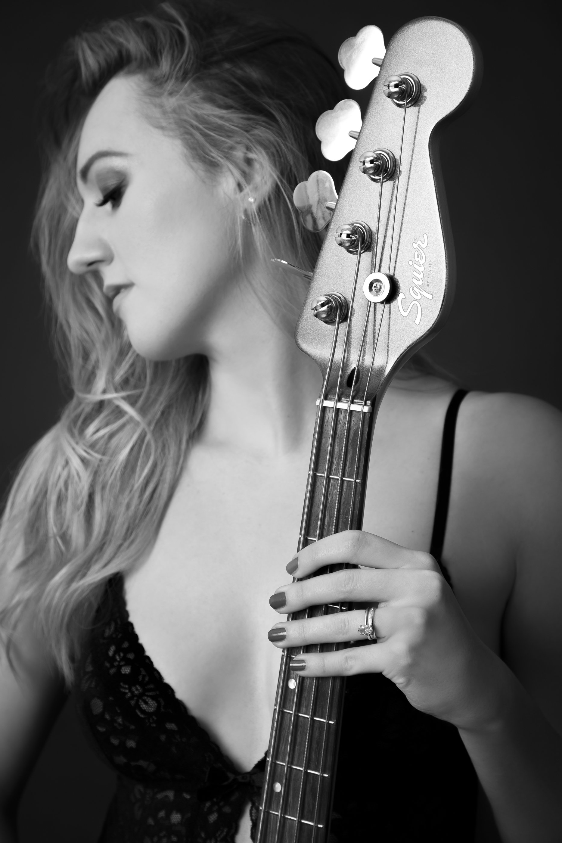 A woman with wavy hair holding a bass guitar in front of her face, wearing a black lace top and a ring on her finger, in black and white.