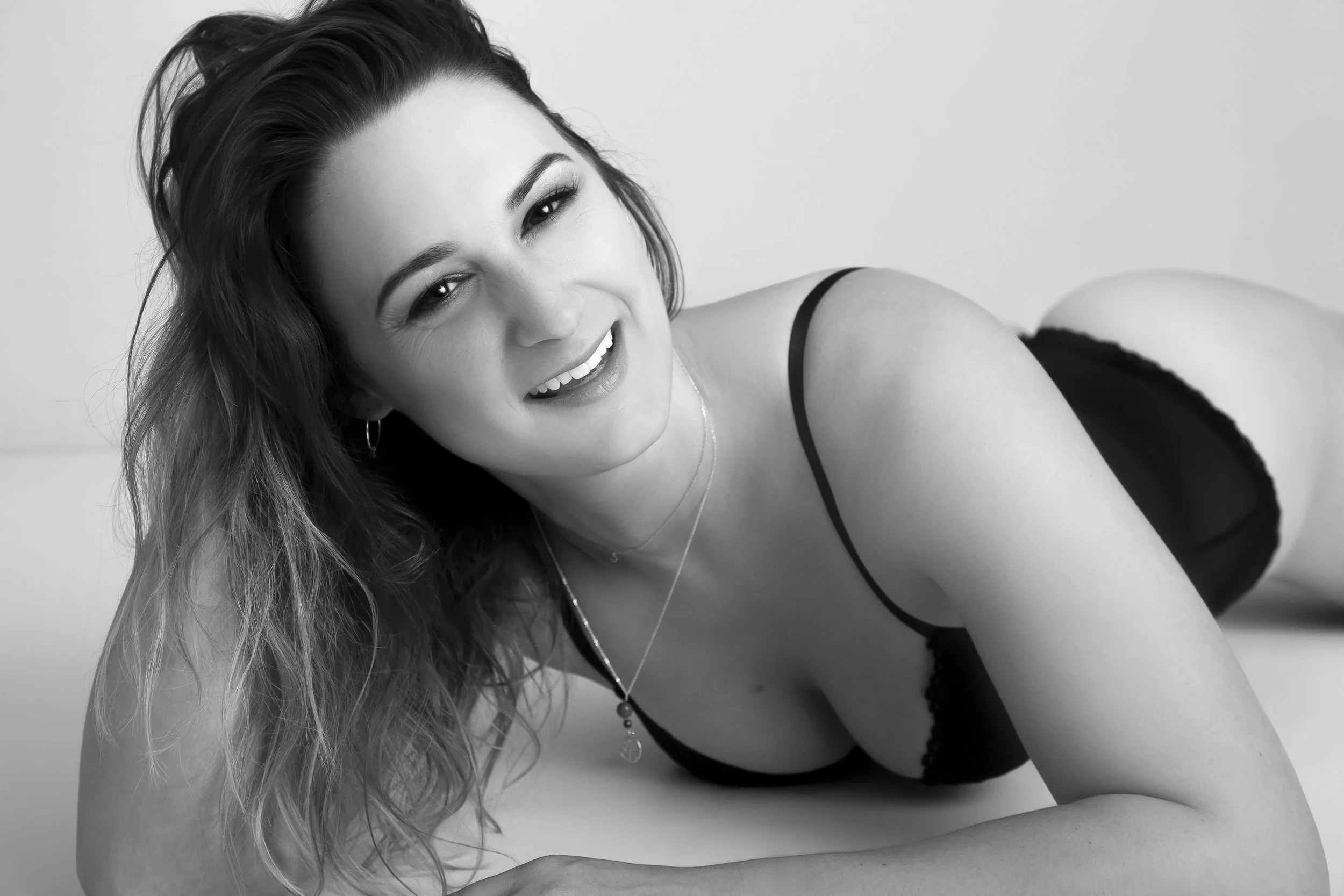Black and white photo of a woman with long, wavy hair lying on her side, smiling, wearing a black bra and jewelry, against a plain background.