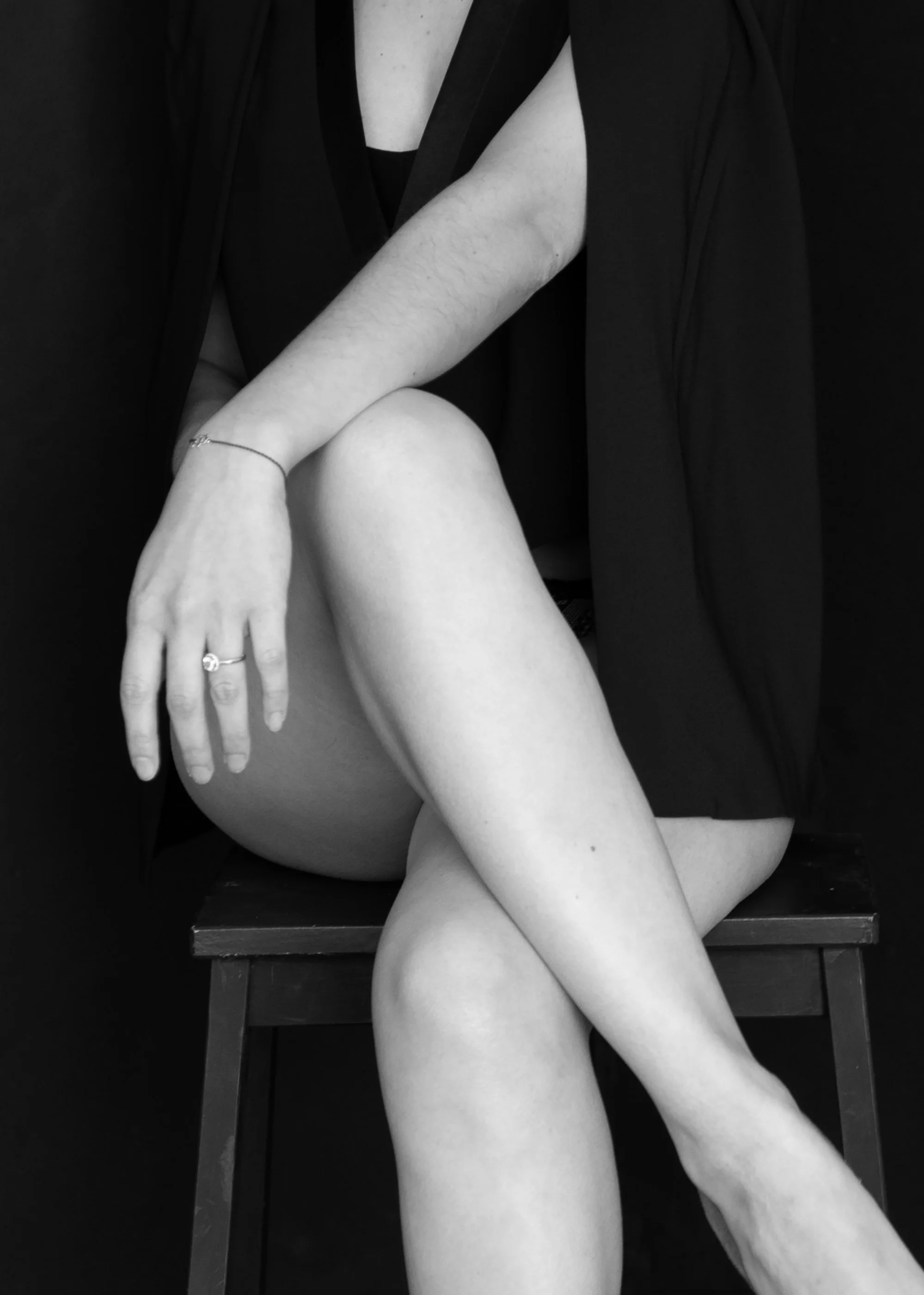 Black and white photo of a woman sitting on a wooden chair with her legs crossed, wearing a black dress and jewelry.
