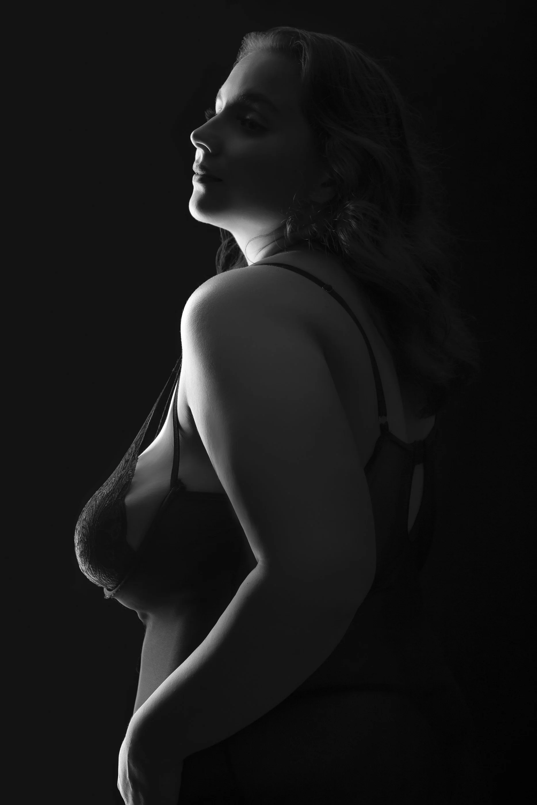 A black-and-white photo of a woman in lingerie, posing with her head turned to the side and her eyes closed, with soft lighting highlighting her profile and shoulder.