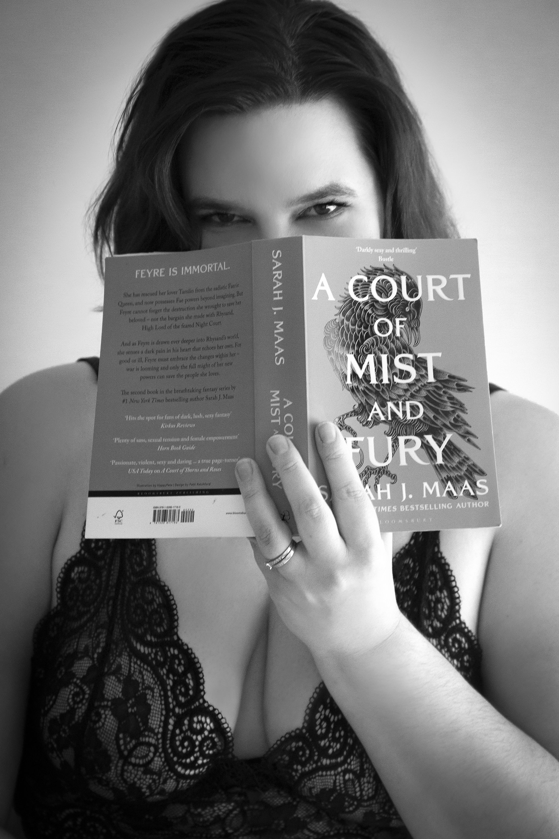 A woman with dark hair wearing a lace top is holding a book titled 'A Court of Mist and Fury' by Sarah J. Maas in front of her face, with her eyes visible over the top of the book.