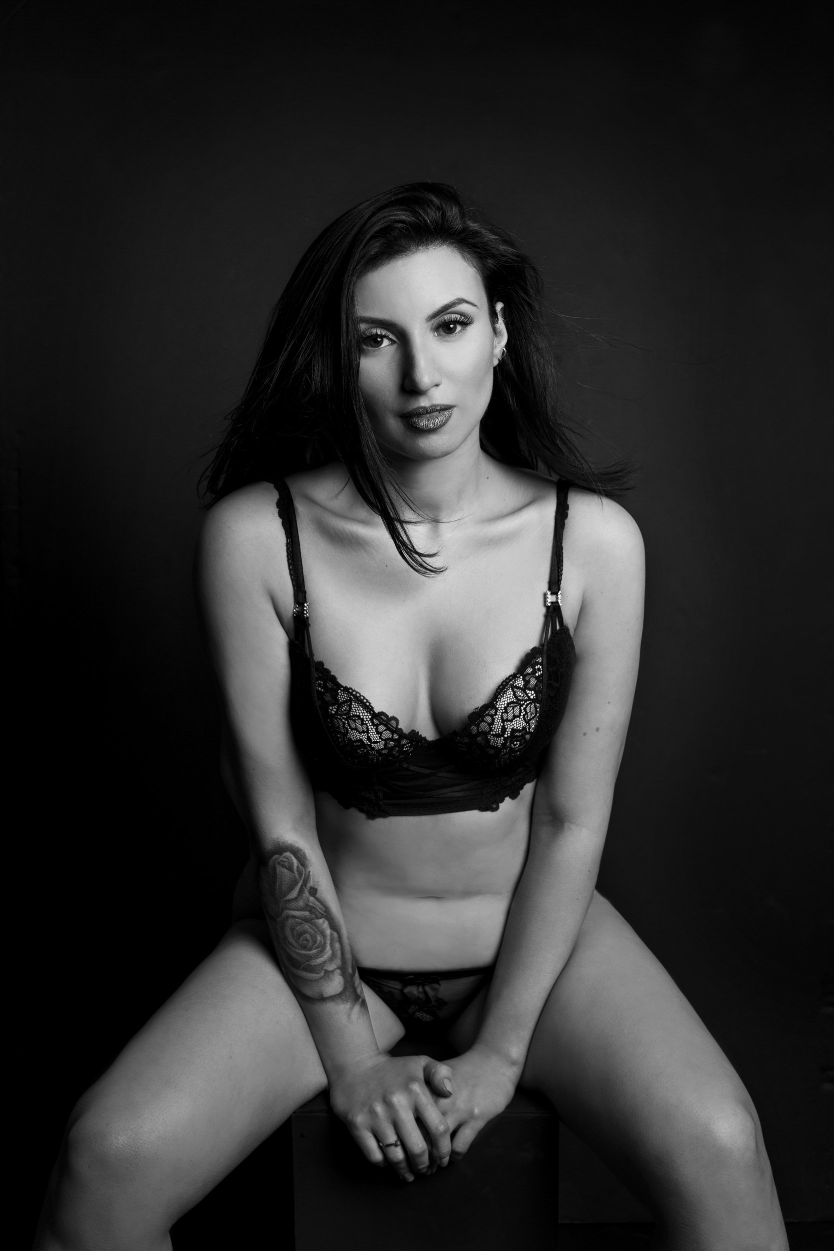 A black-and-white portrait of a woman with dark hair, wearing lace lingerie, sitting with her legs apart and hands clasped together between her legs against a dark background.