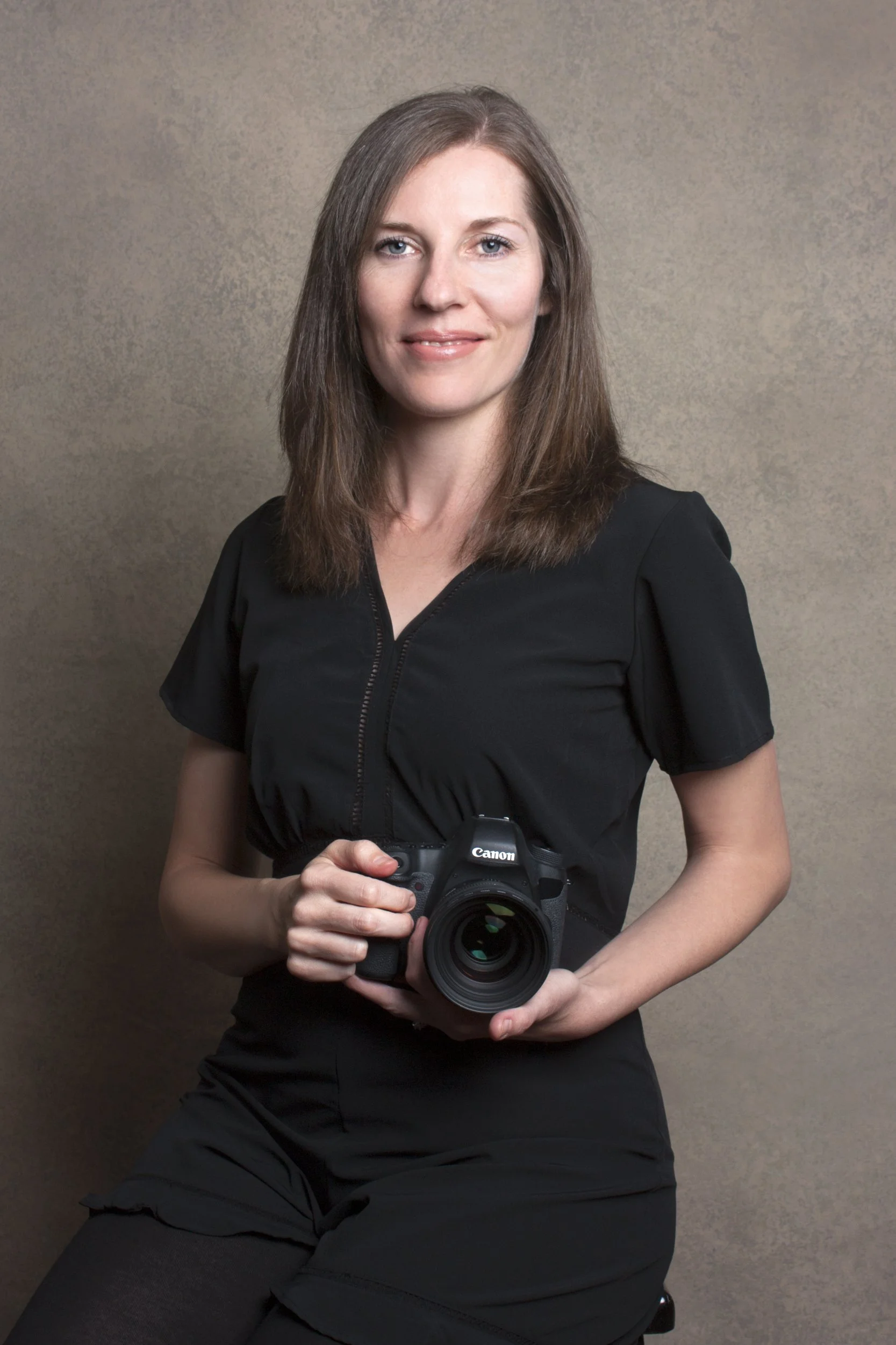 A woman with brown hair holding a Canon camera, wearing a black top, standing against a plain background.