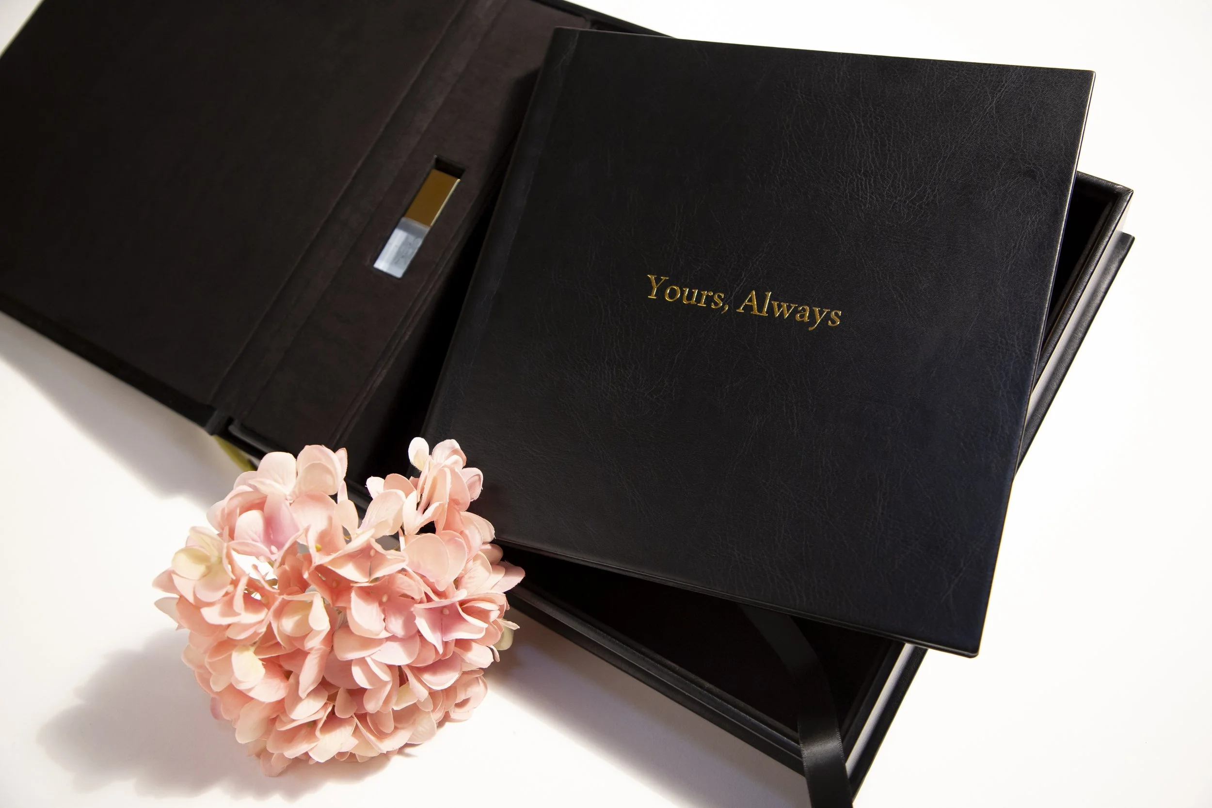 A black leather photo album with gold lettering that says 'Yours, Always', a pink hydrangea flower, and a black presentation box.