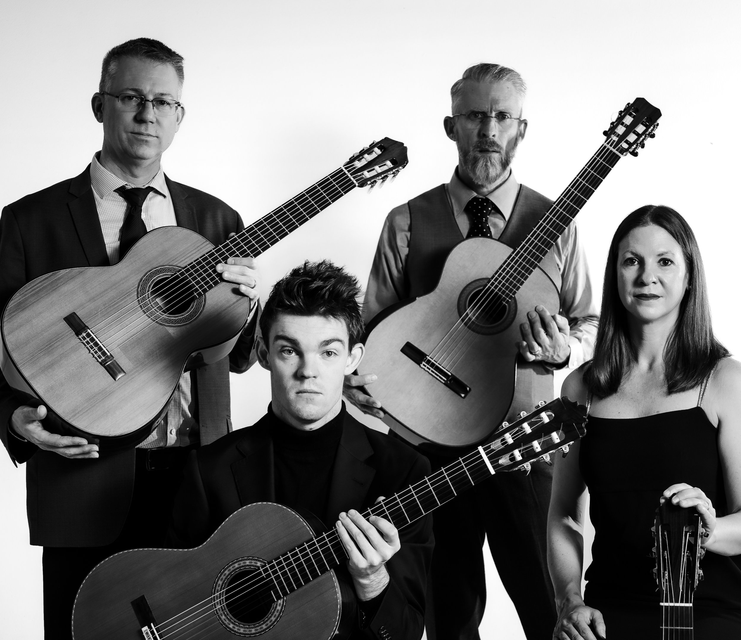 Black and white photo of four people holding acoustic guitars, dressed in formal attire, posing against a plain background.