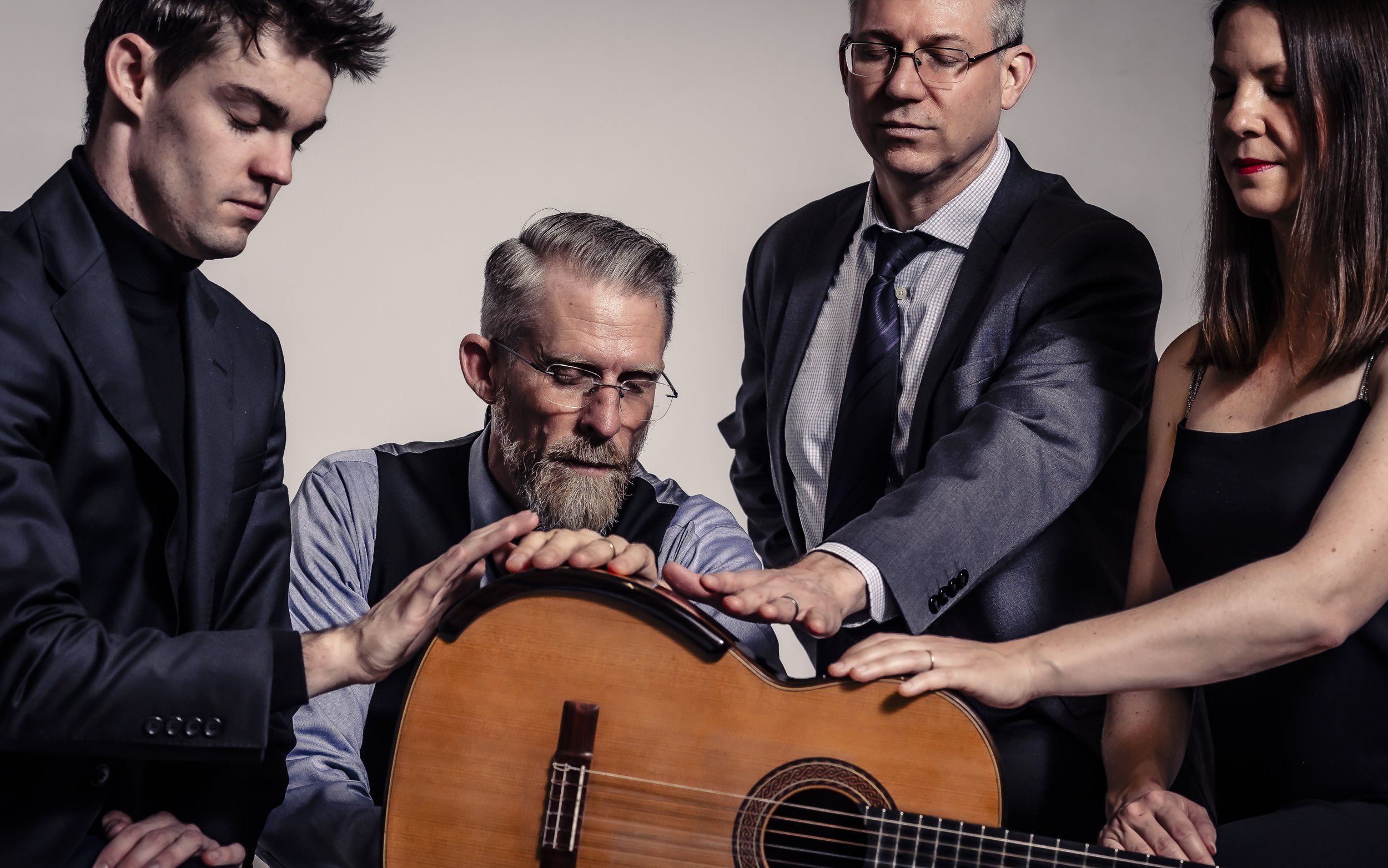 Four people with closed eyes touching a guitar with their hands, wearing formal attire, against a plain background.