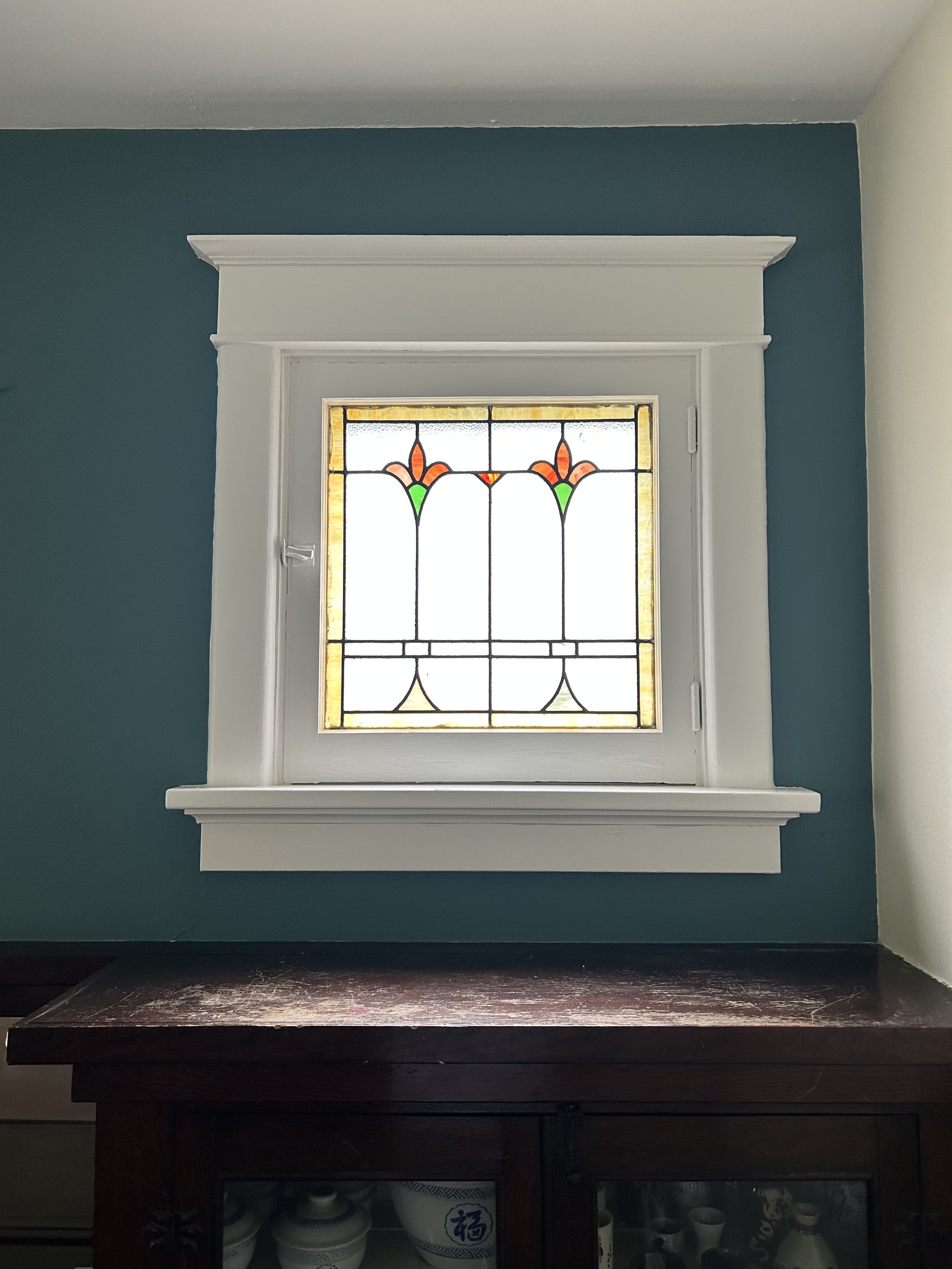 Decorative stained glass window with floral patterns in orange and green, framed by white trim, set against a teal wall above a dark wooden cabinet.