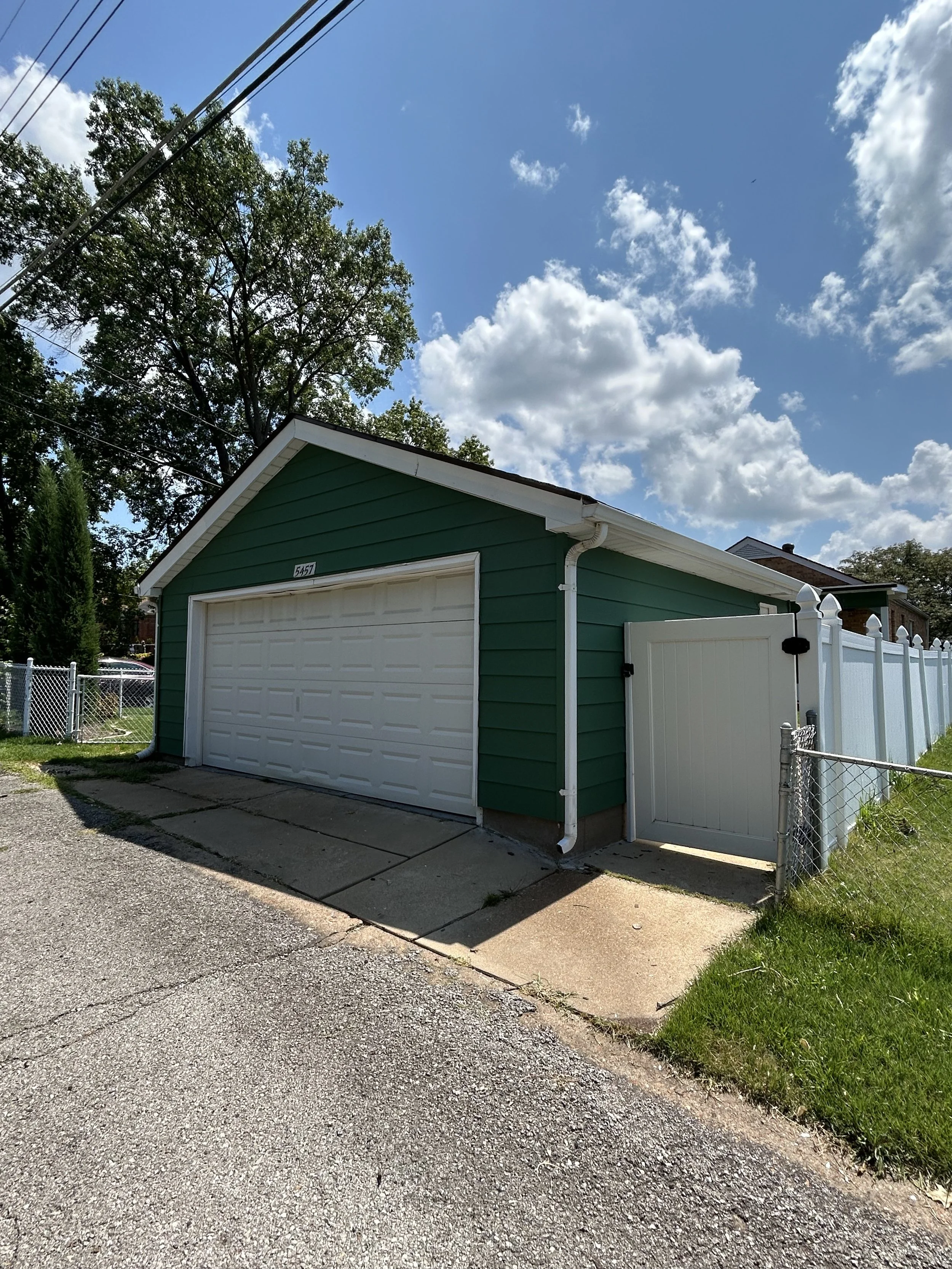 A green garage with a white roll-up door, a small side door, and a white picket fence gate, with a concrete driveway and sidewalk, under a partly cloudy sky.