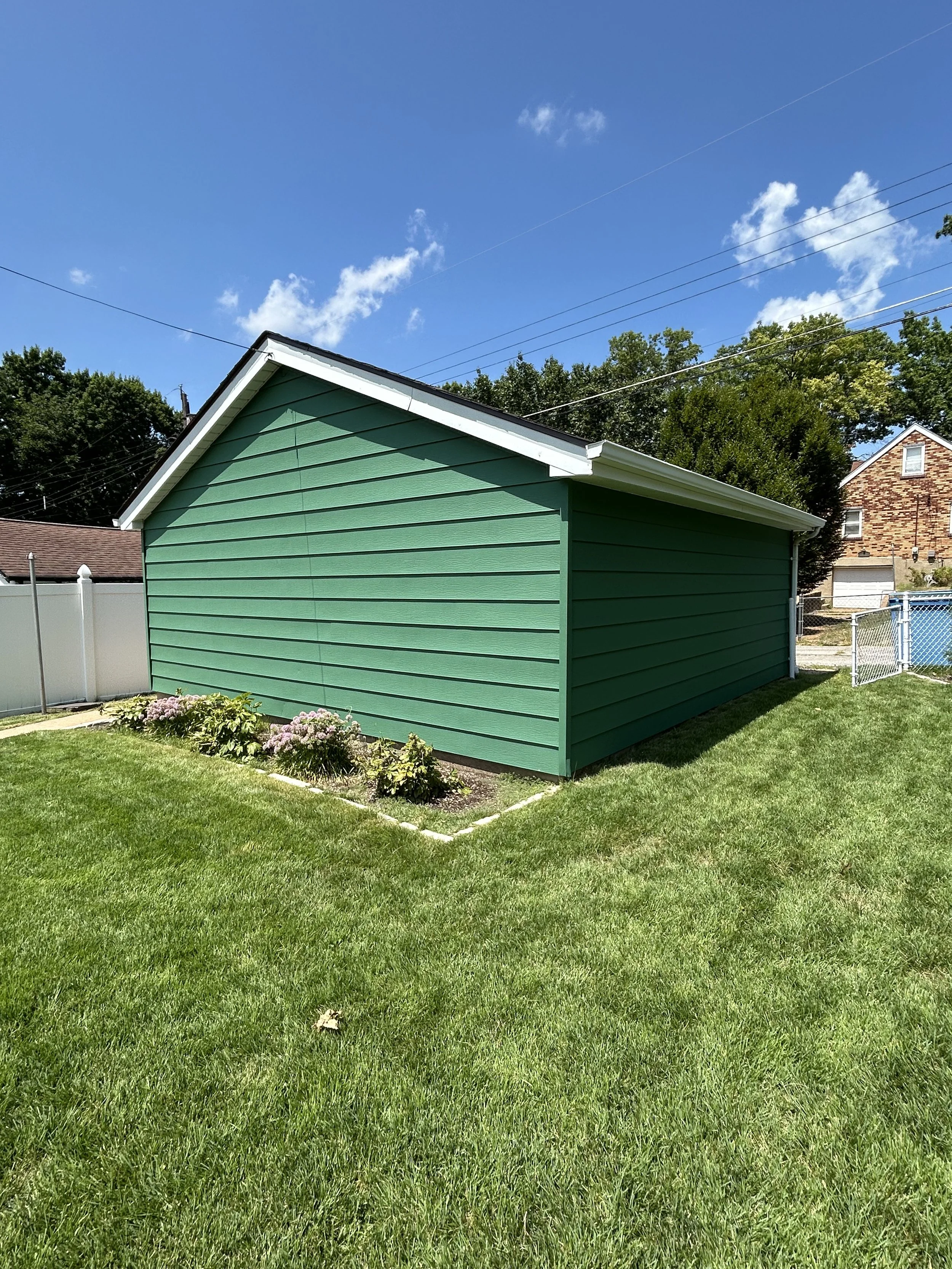 A green backyard shed with white trim in a grassy yard under a blue sky with scattered clouds.