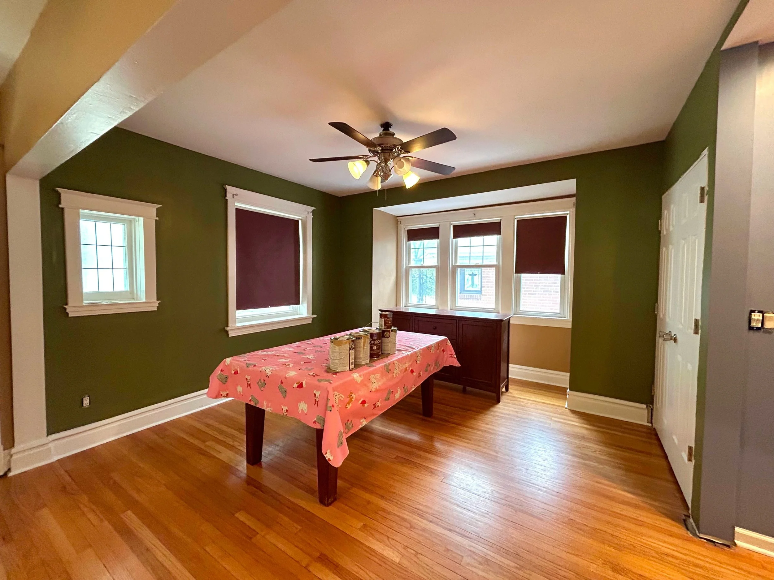 Dining room with green walls, hardwood floors, and multiple windows with white trim. There is a table covered with a pink tablecloth decorated with Christmas-themed designs, and a ceiling fan with lights.