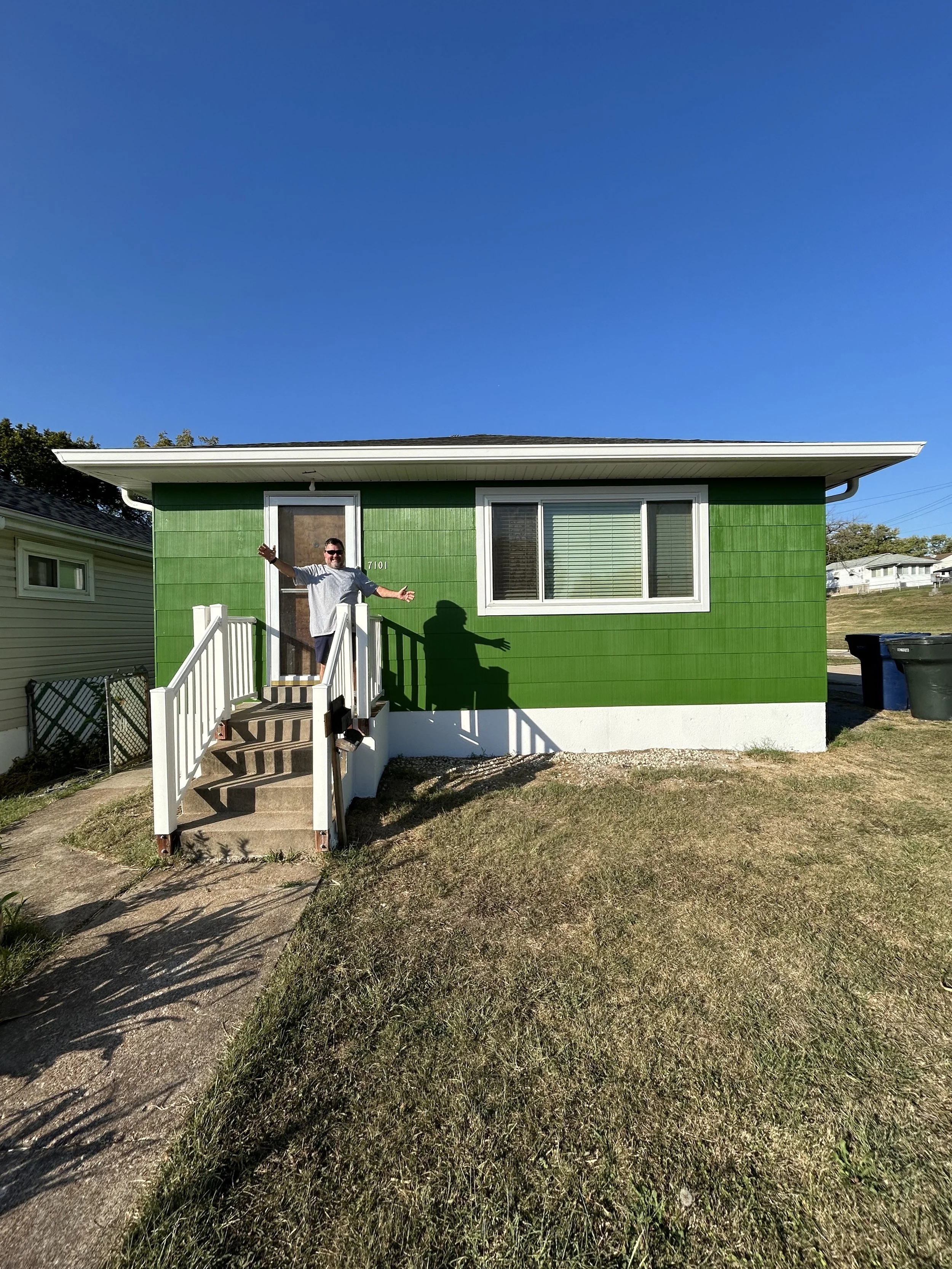 Man standing on the front steps of a green house with a white railing, raising his arms, under a clear blue sky.