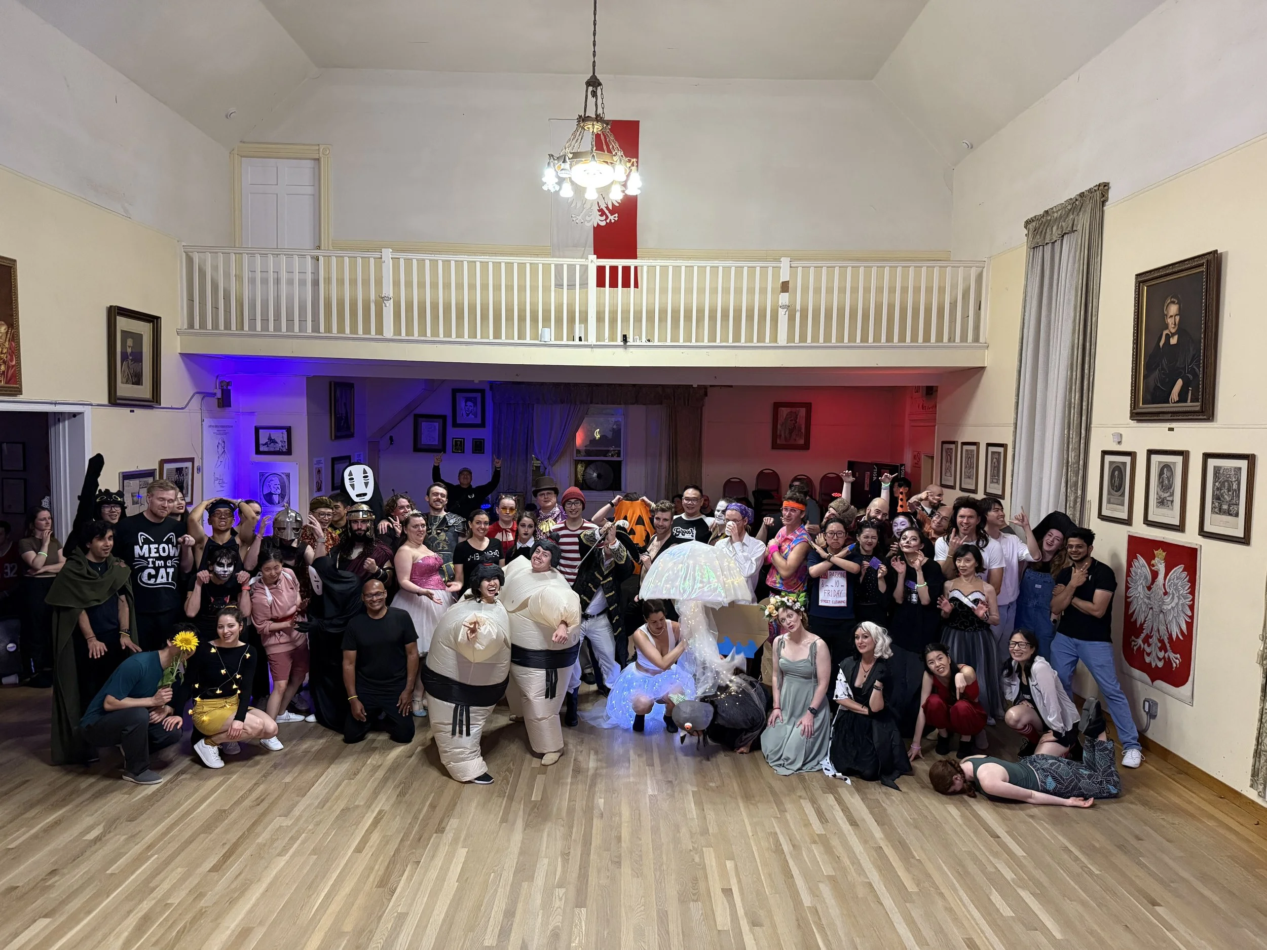 A large group of people in costume gathering in a spacious room with framed pictures on the walls, some people holding props and making gestures, with a band or performers at the front.