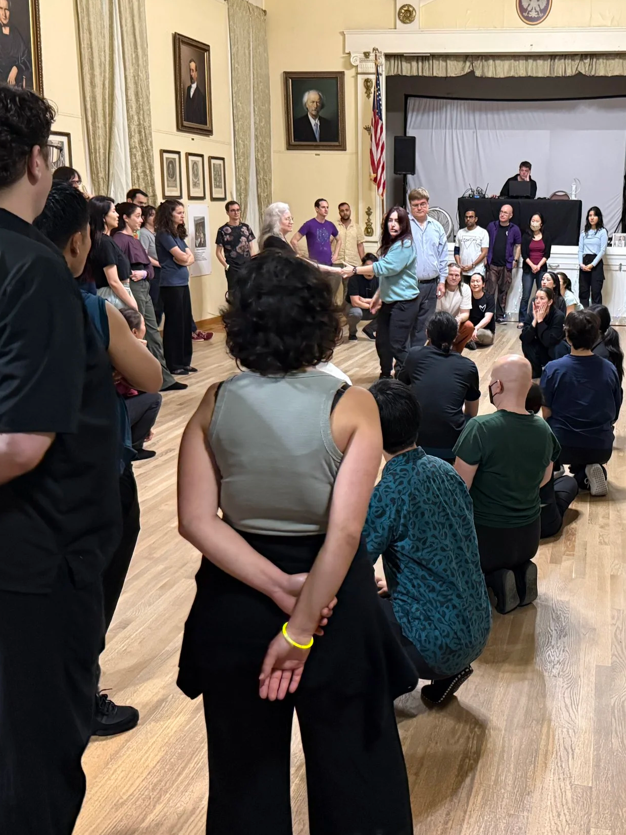 A group of people gathered in a room with framed portraits on the walls, some seated on the floor and others standing, as two individuals dance in the center. The setting appears to be an event or workshop in a historic or official building.