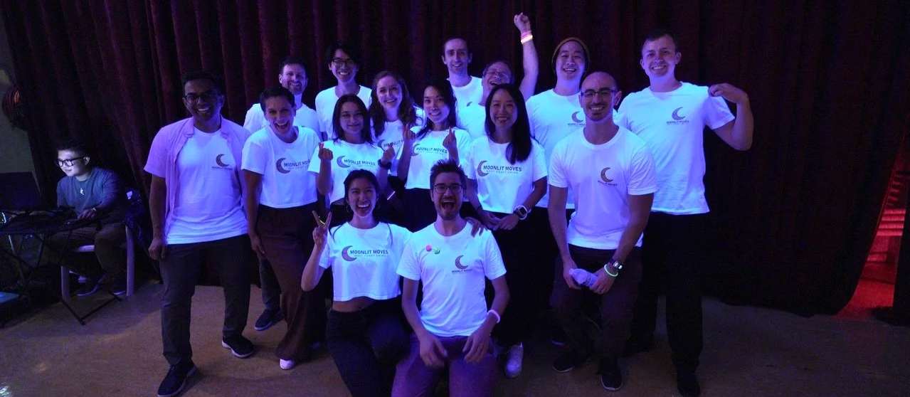 Group of people on stage wearing matching white T-shirts with a moon logo, posing for a photo and smiling, some making peace signs, with purple lighting and dark curtains in the background.