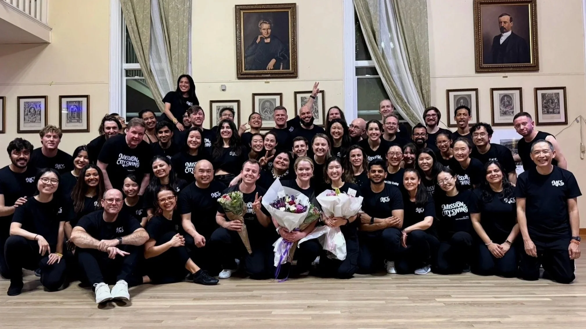 A large group of smiling people posing together in a room with framed portraits on the wall. Two women in the front hold bouquets of flowers, and most individuals are wearing matching black t-shirts with the logo 'MISSION CITY SWING'.