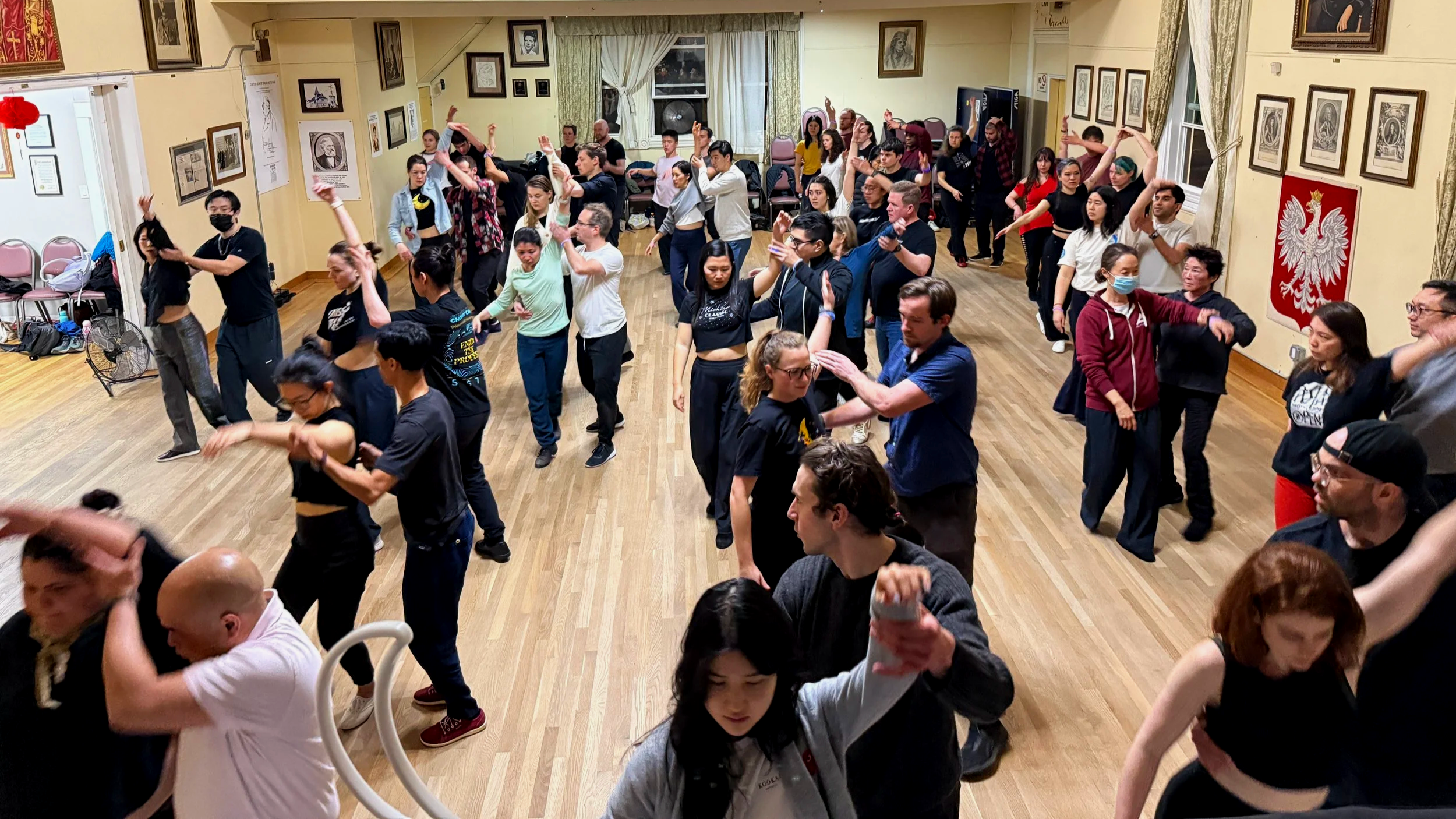 A group of people dancing together in a room with wooden floors and walls decorated with framed photos and artwork; some wearing masks, others not, engaged in a social dance event.