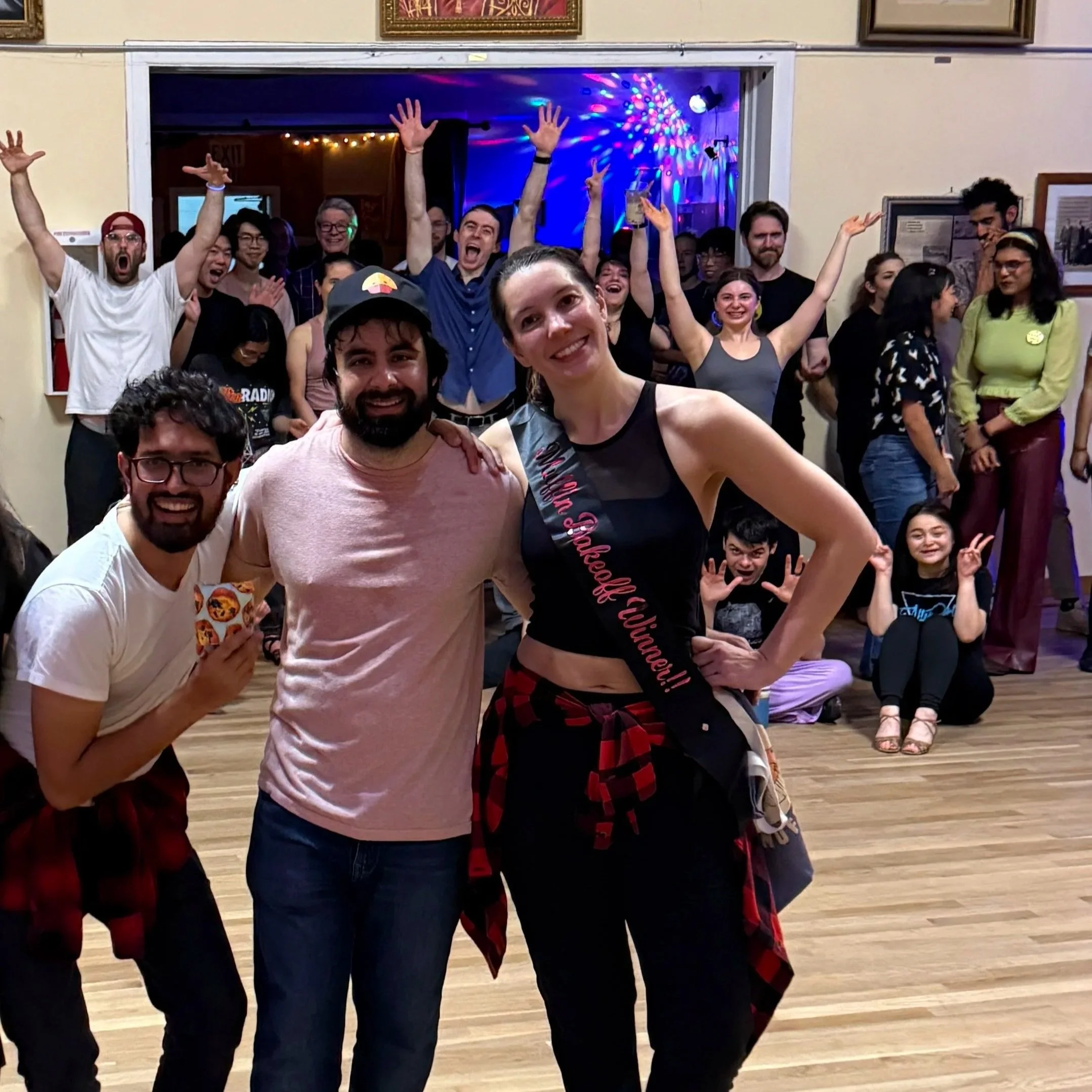 A group of smiling young adults dancing and celebrating at a lively party or social gathering indoors with colorful lights and a wooden floor.