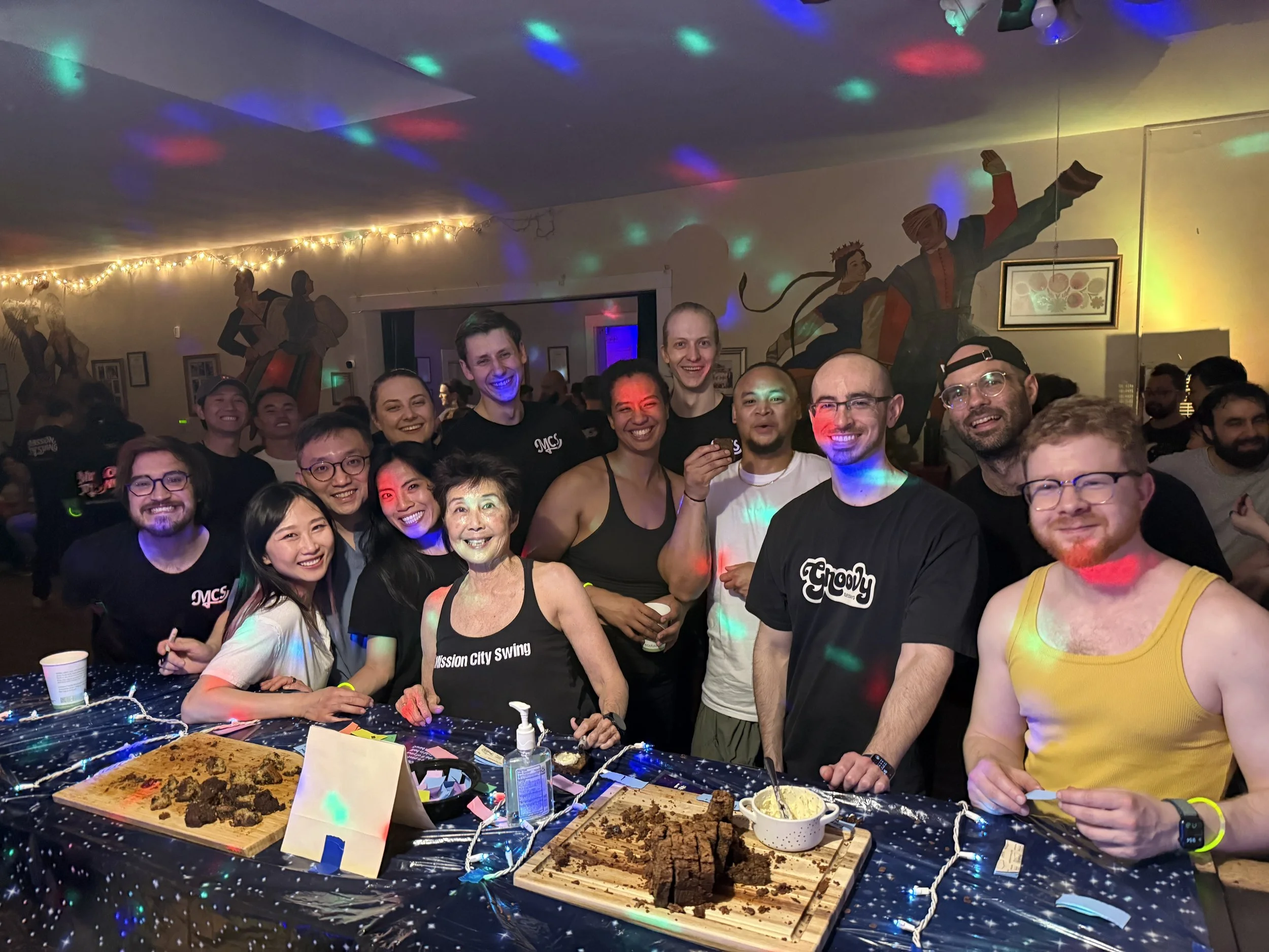 A group of smiling people gathered around a table at a party, with some holding drinks. The table has cake, plates, and party decorations. In the background, there is a colorful mural and string lights on the wall, and the room is lit with multicolored disco lights.