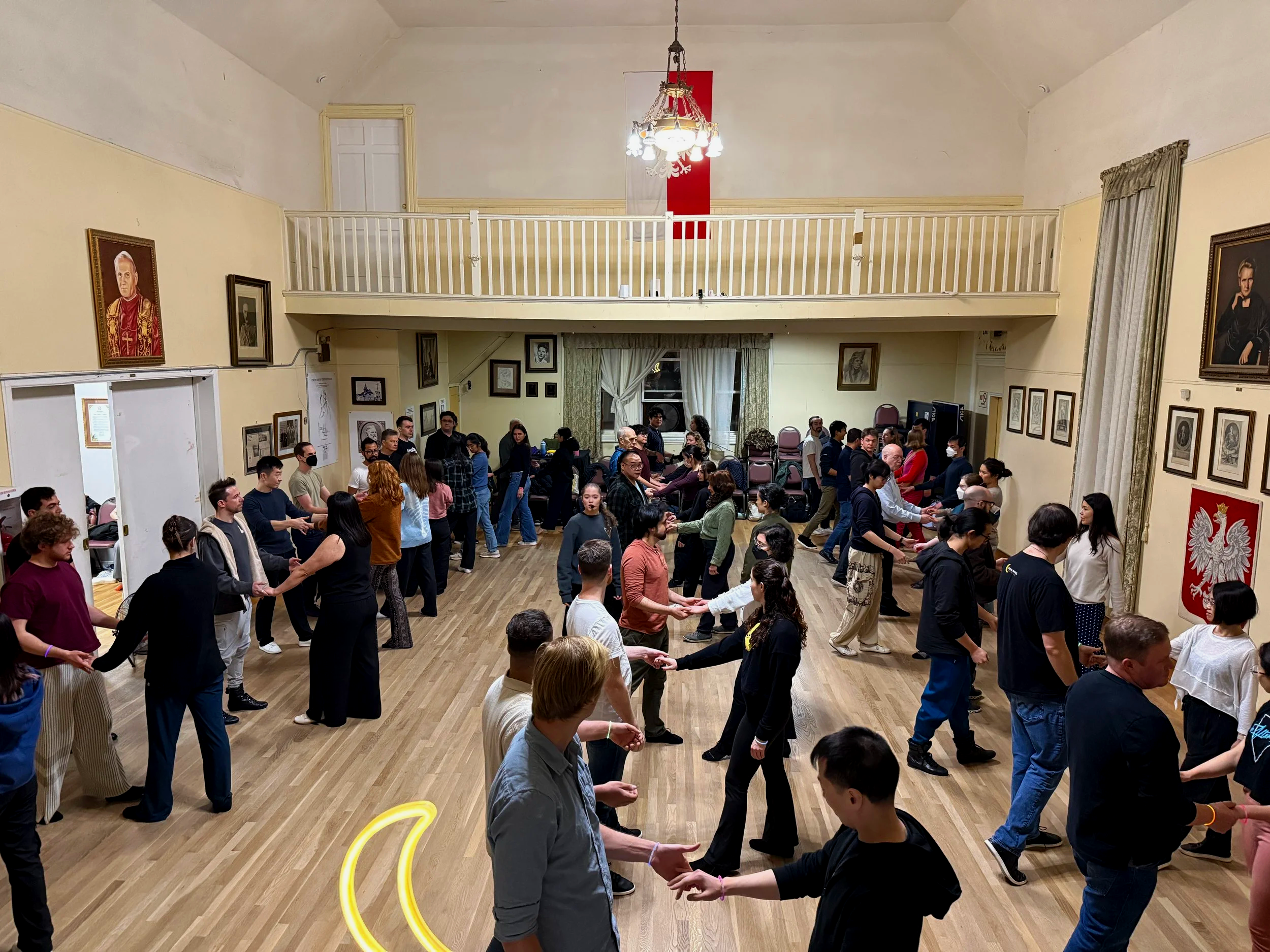 People holding hands in a circle, dancing or participating in a group activity inside a large room with portraits on the walls and a red and white flag at the back.