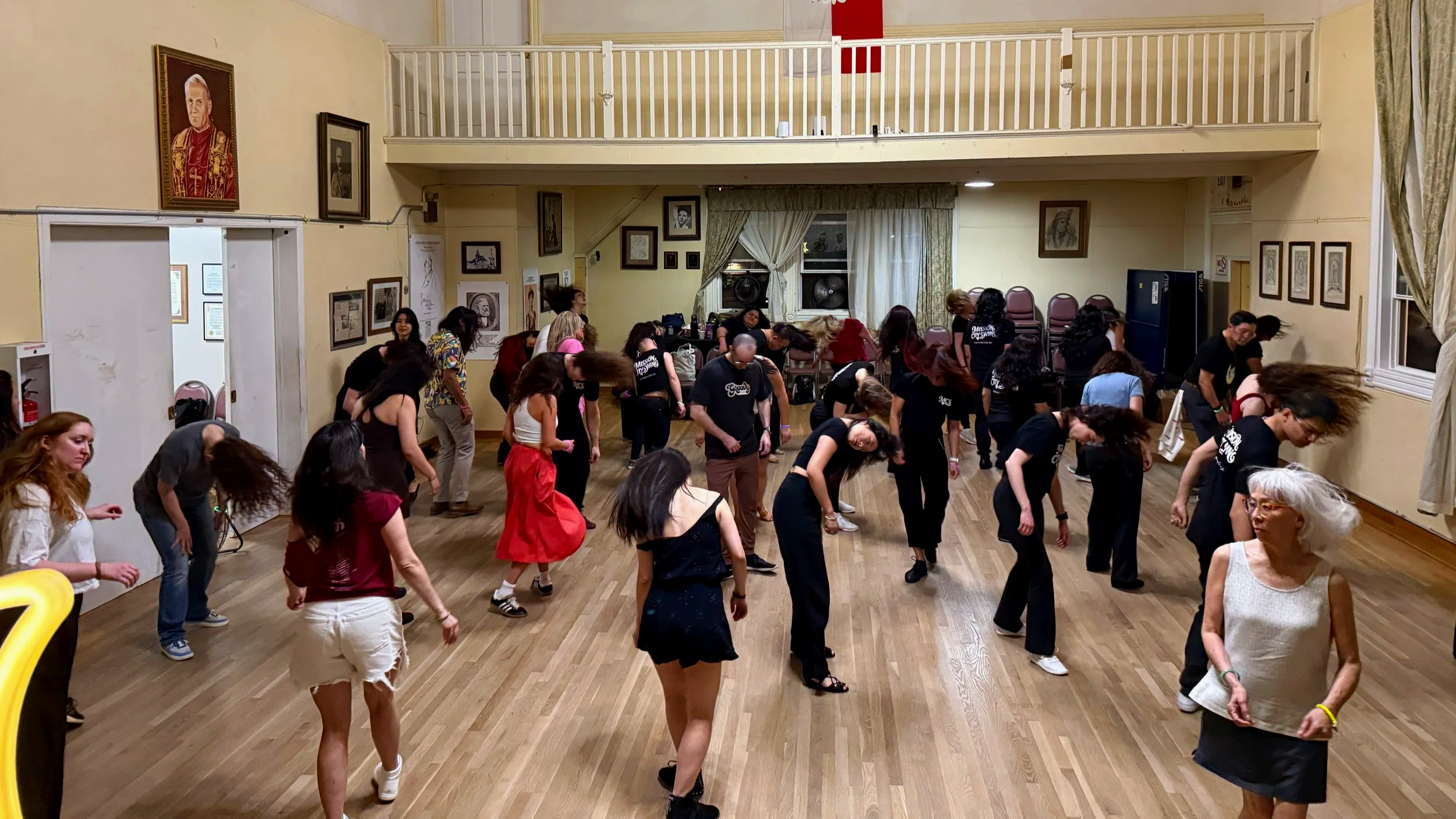 Group of people participating in a dance class or social dance event in a spacious room with wooden floors, framed pictures on the walls, and curtains on windows.