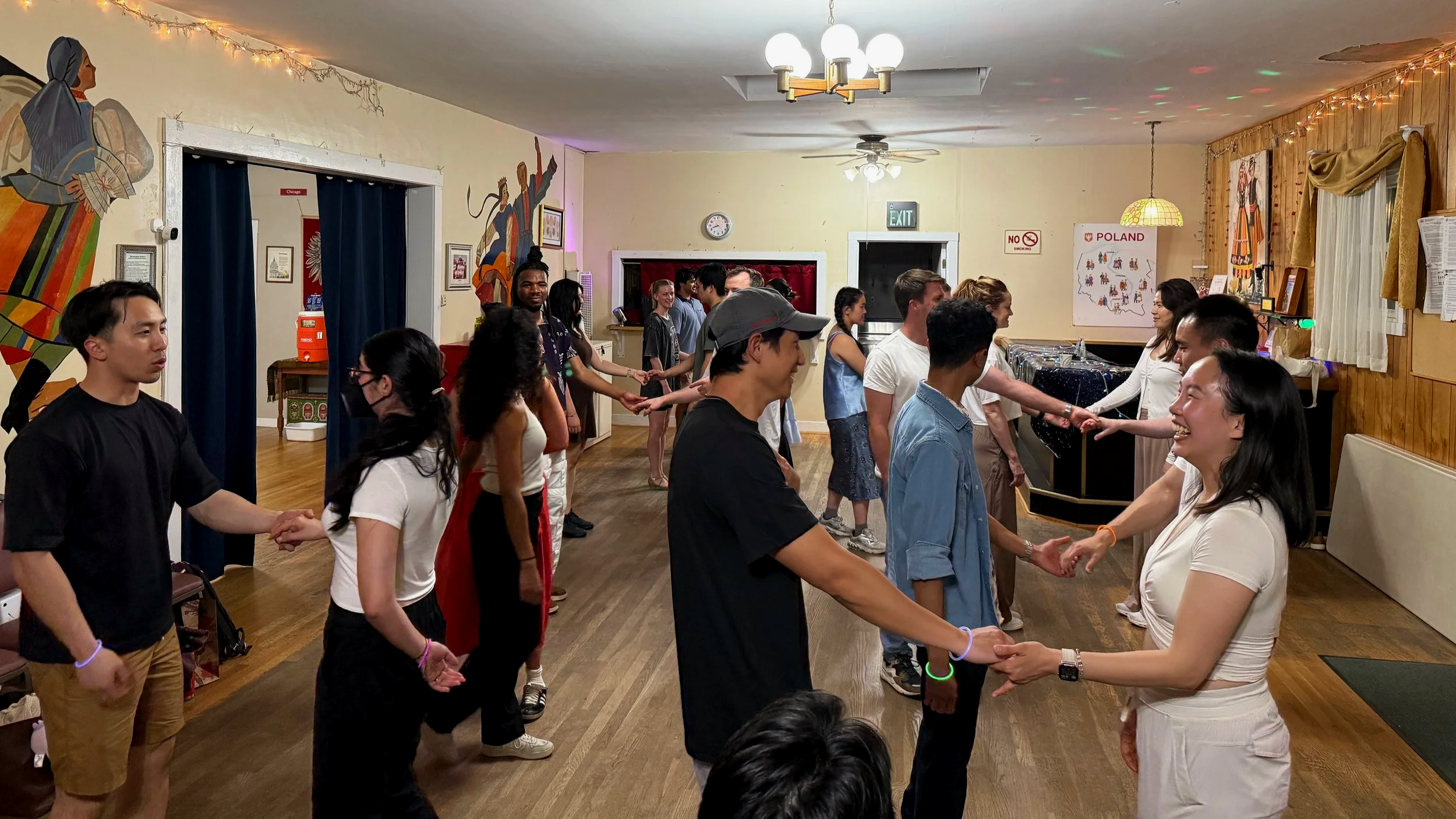 People taking part in a dance or circle game in a cozy decorated room, with some holding hands and smiling, while others watch and participate. The room has wooden walls, festive lights, and cultural artwork.