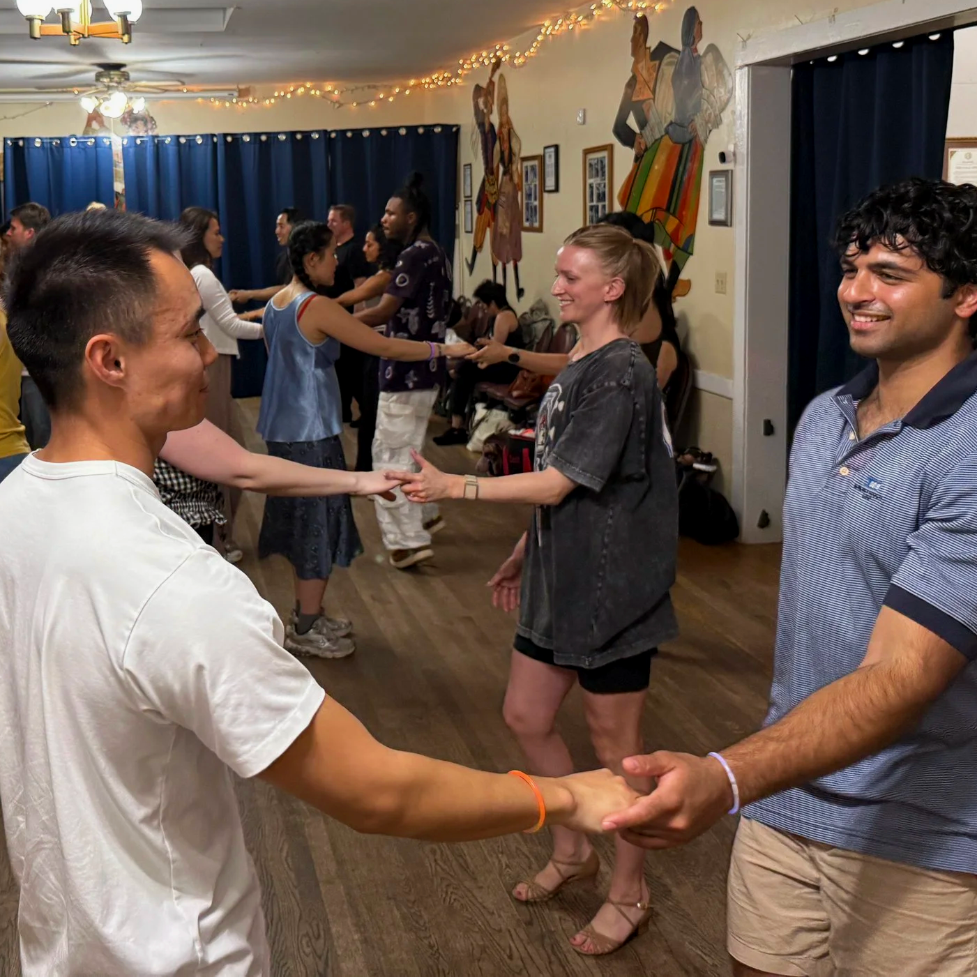People dancing in a circle, holding hands at a social gathering or party, with decorative murals on the wall and string lights overhead.