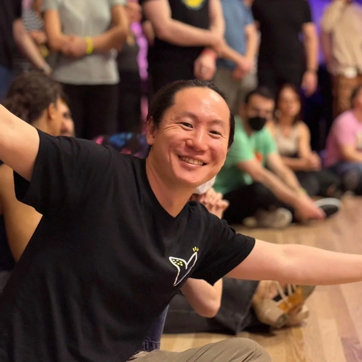 A smiling man with black hair taking a selfie at a social gathering, with a group of people sitting and standing in the background.