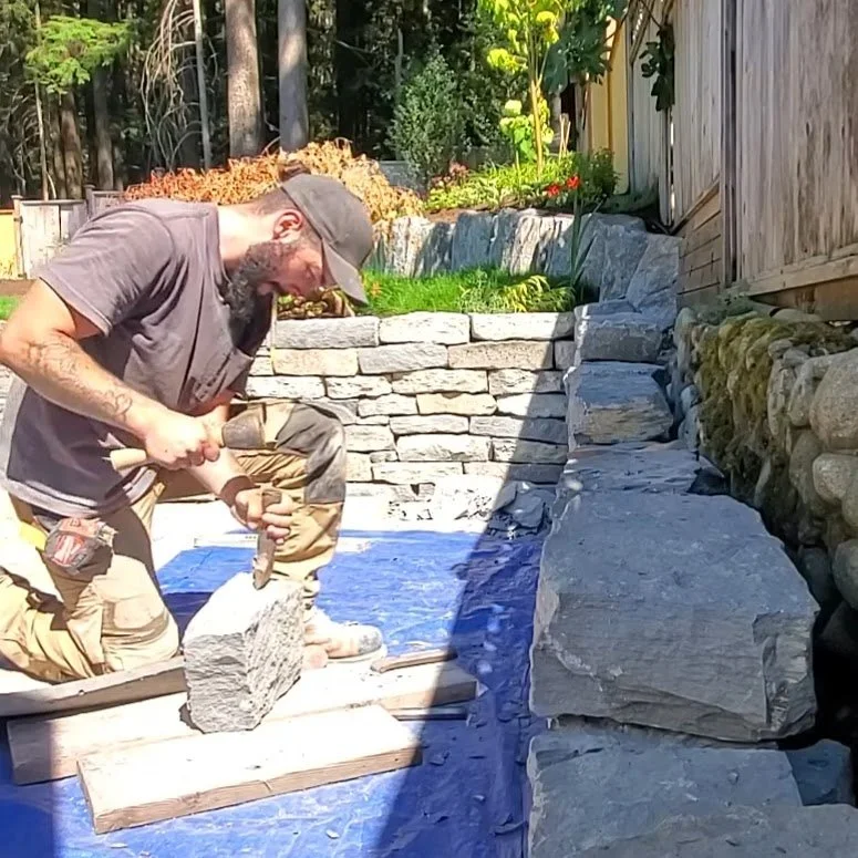Man with a beard and cap working with a hammer on a stone block in a backyard with stone walls and plants.