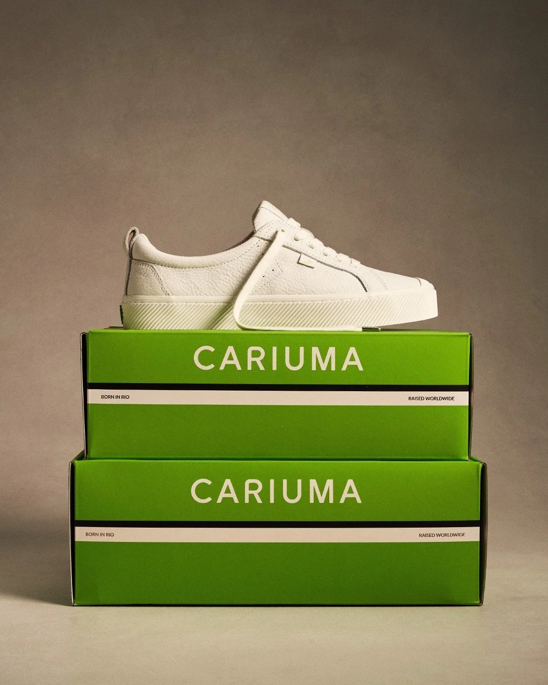 A white sneaker resting on top of two green shoeboxes labeled 'CARIUMA', with the shoe's laces undone, in a neutral background.