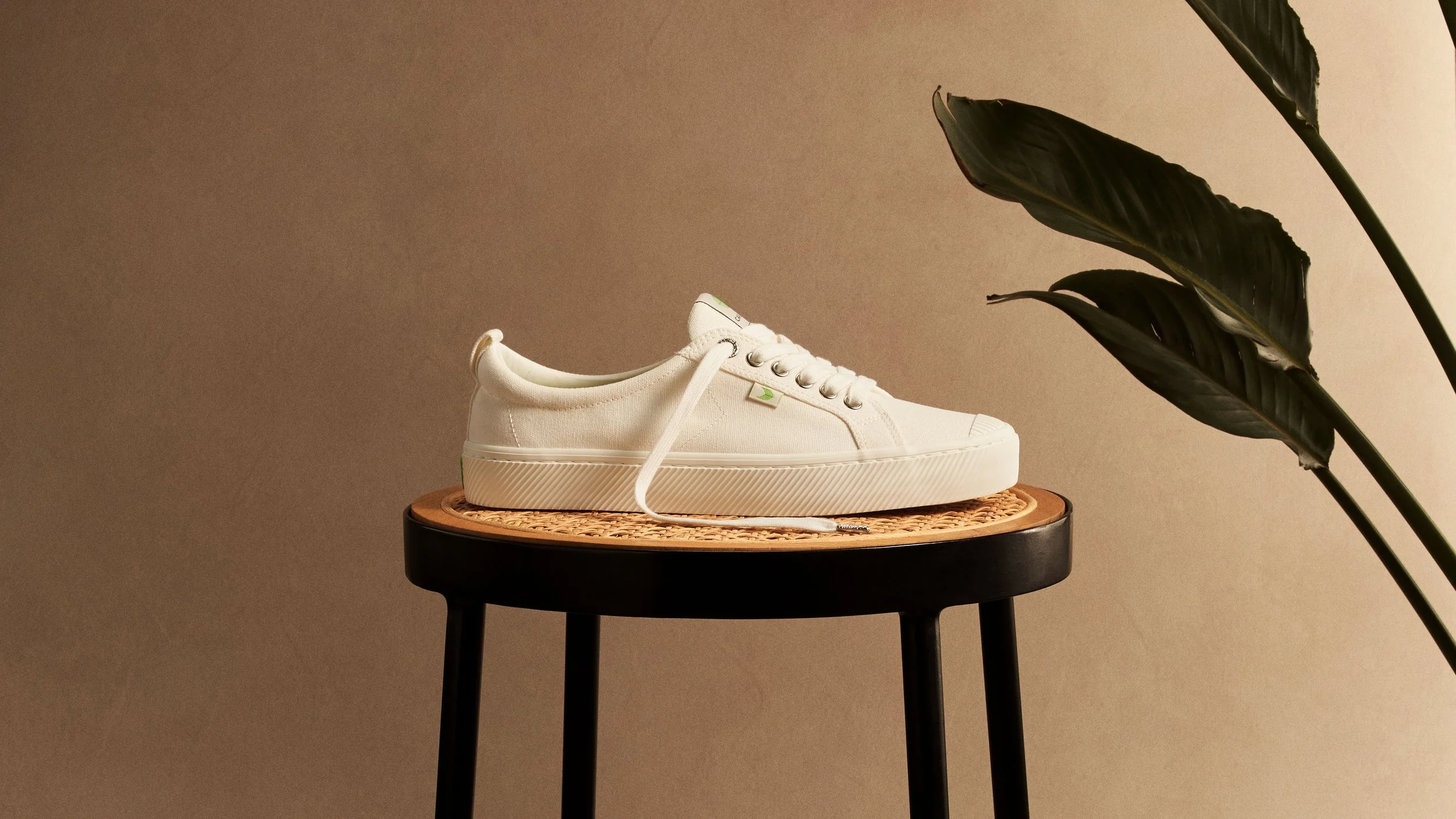 A white sneaker with a textured rubber sole on a black and natural wood table, with large dark green leaves in the background.