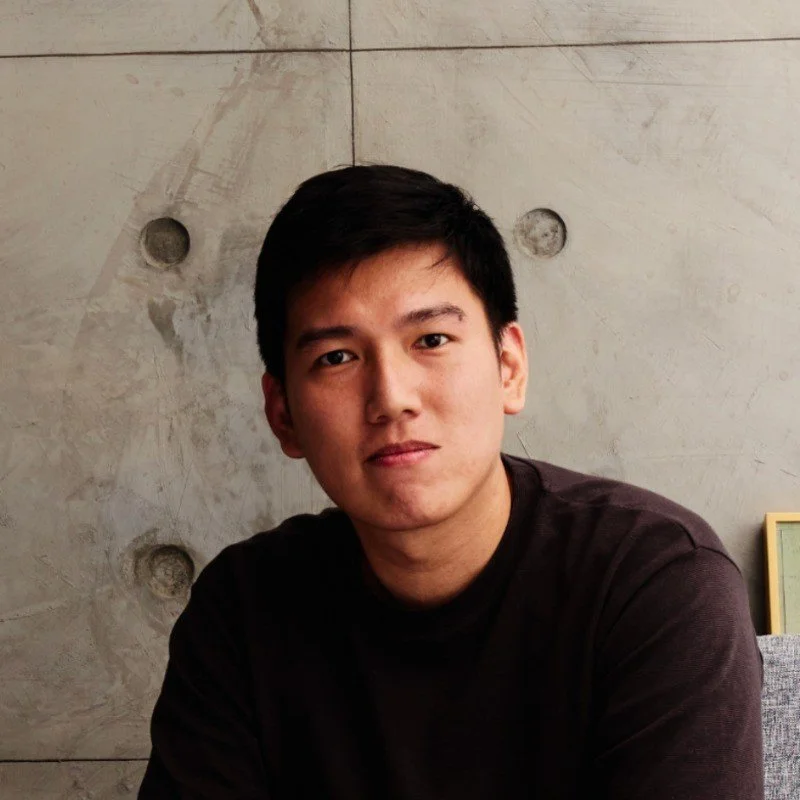 A young man with black hair, wearing a black shirt, sitting in front of a light-colored wall with circular indentations.