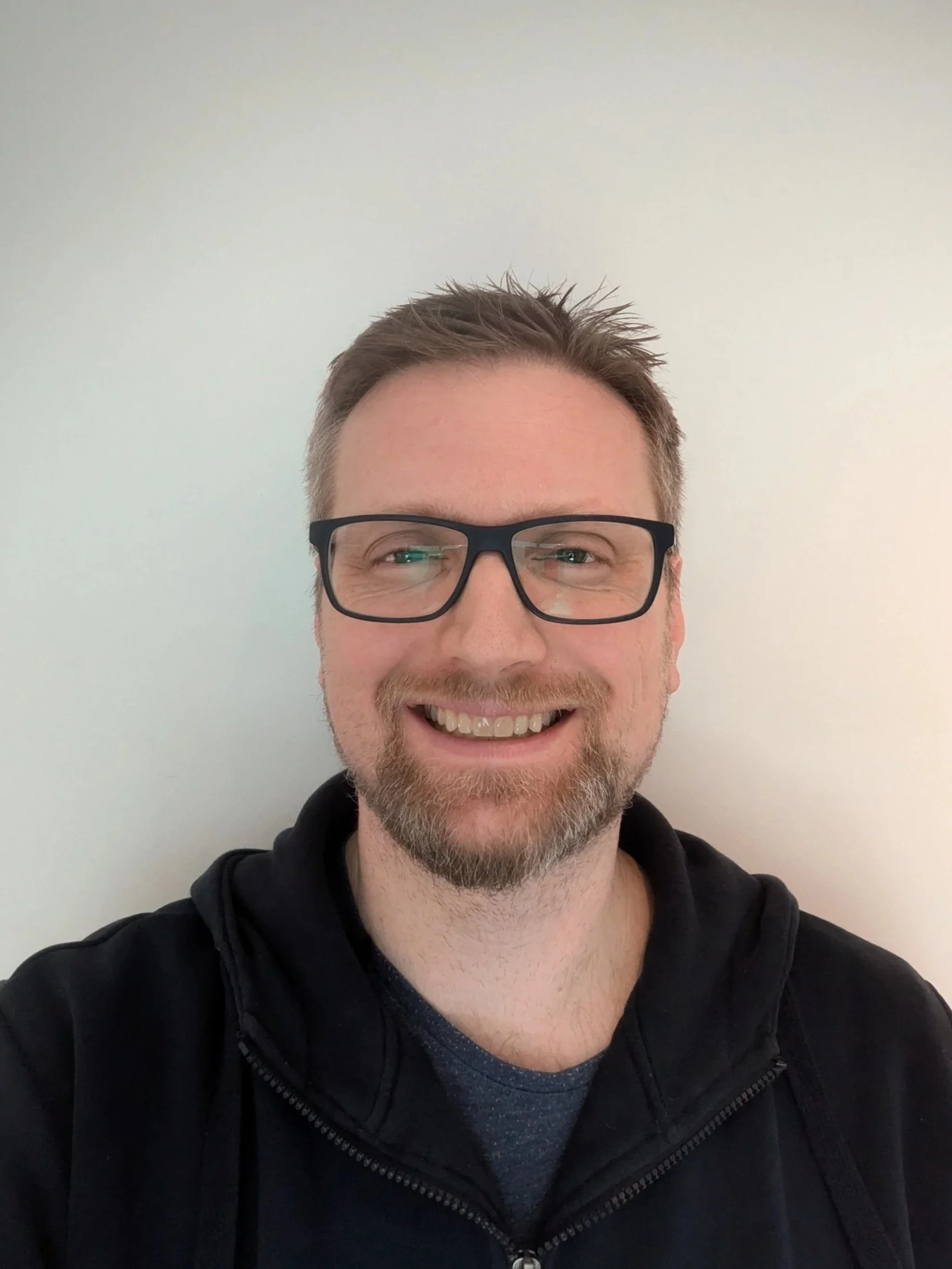 Nærbilde av en mann med brunt hår, skjegg og briller, mot en lys bakgrunn.