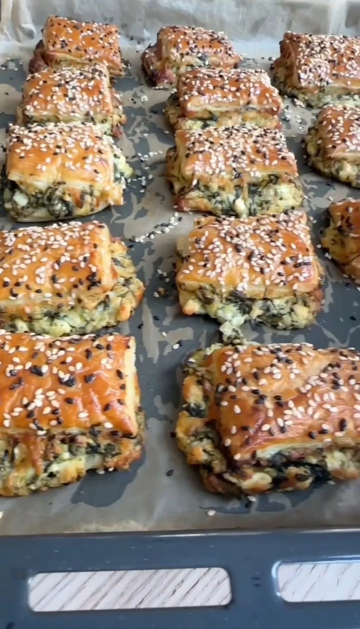 Baked spinach and cheese puff pastry squares topped with white sesame seeds and black sesame seeds on a baking sheet.