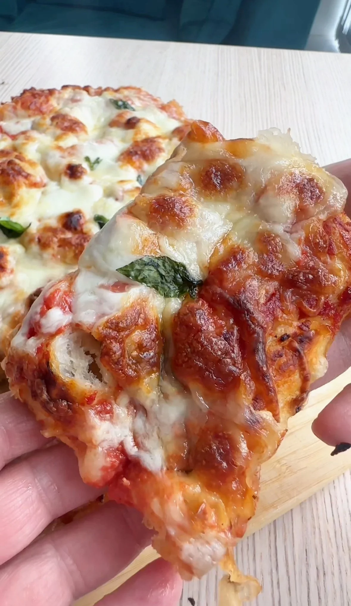 Close-up of person holding a slice of cheese and tomato pizza with basil on a wooden board, with another whole cheese and tomato pizza in the background.