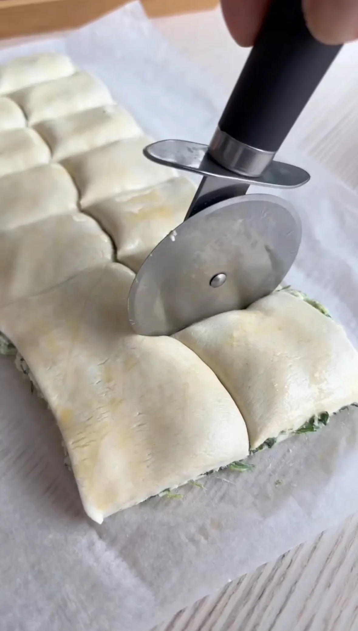 A pizza cutter slicing through a rolled-out pizza dough with green vegetables visible underneath.