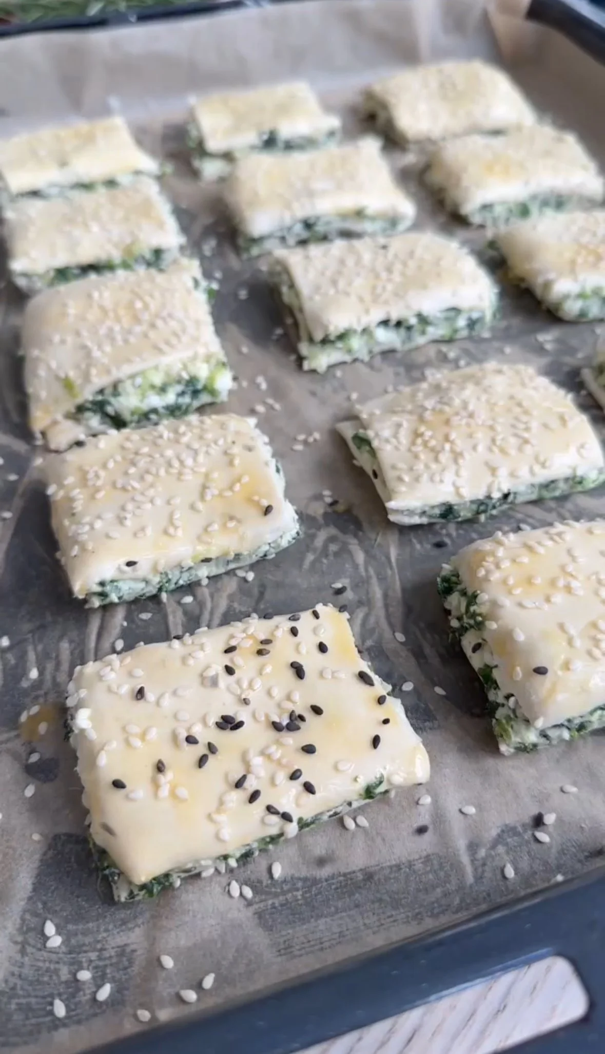 Unbaked spinach and cheese pastries topped with sesame seeds on a baking sheet lined with parchment paper.