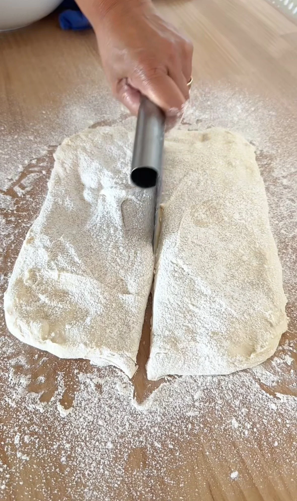 A person cutting dough in half with a metal dough cutter on a floured wooden surface.