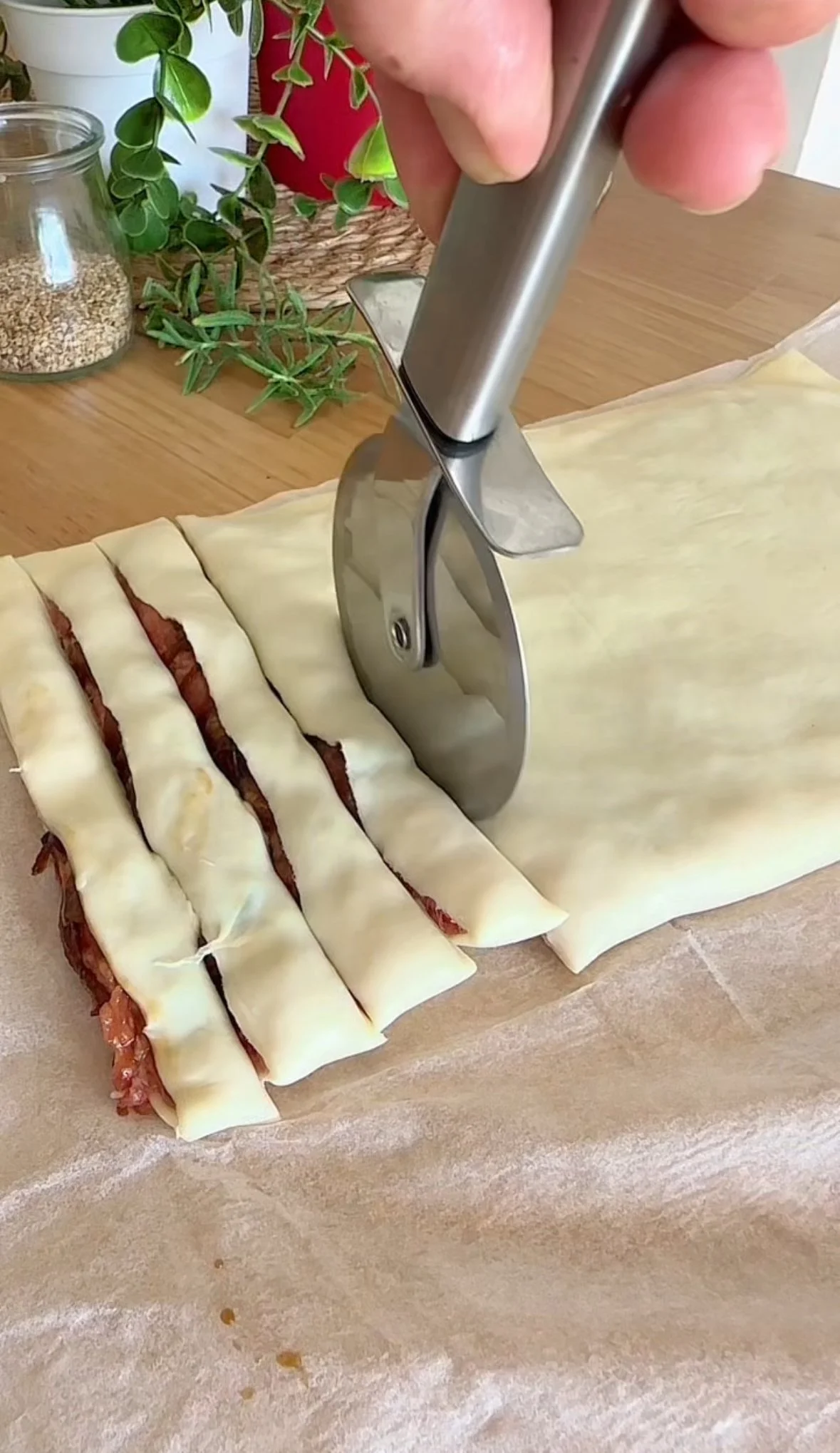 Close-up of a hand using a pizza cutter to slice a rolled-out dough with filling, on parchment paper, with potted plants and spices in the background.