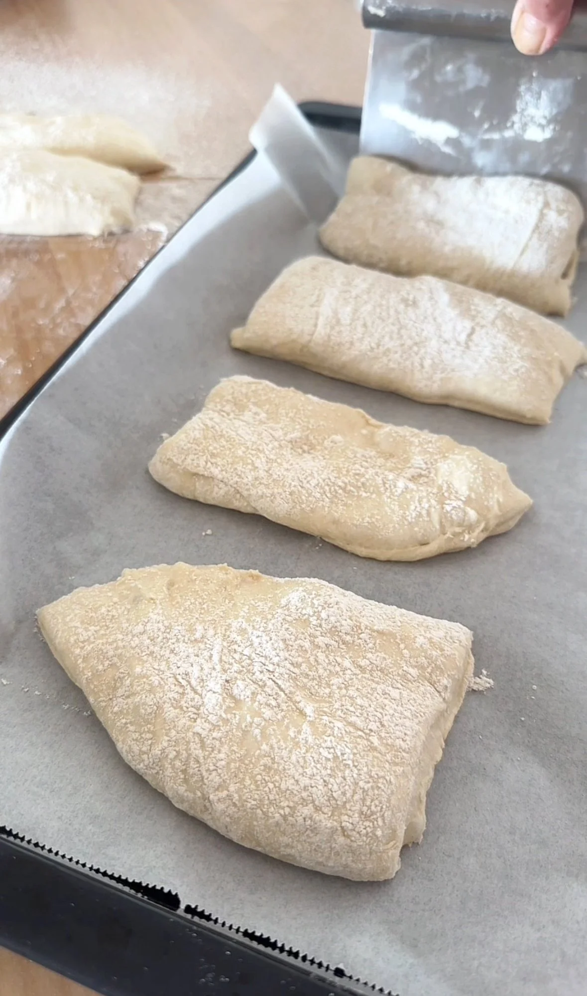 Four pieces of dough are rolled and sprinkled with flour on a baking sheet, with someone cutting the dough into smaller sections.