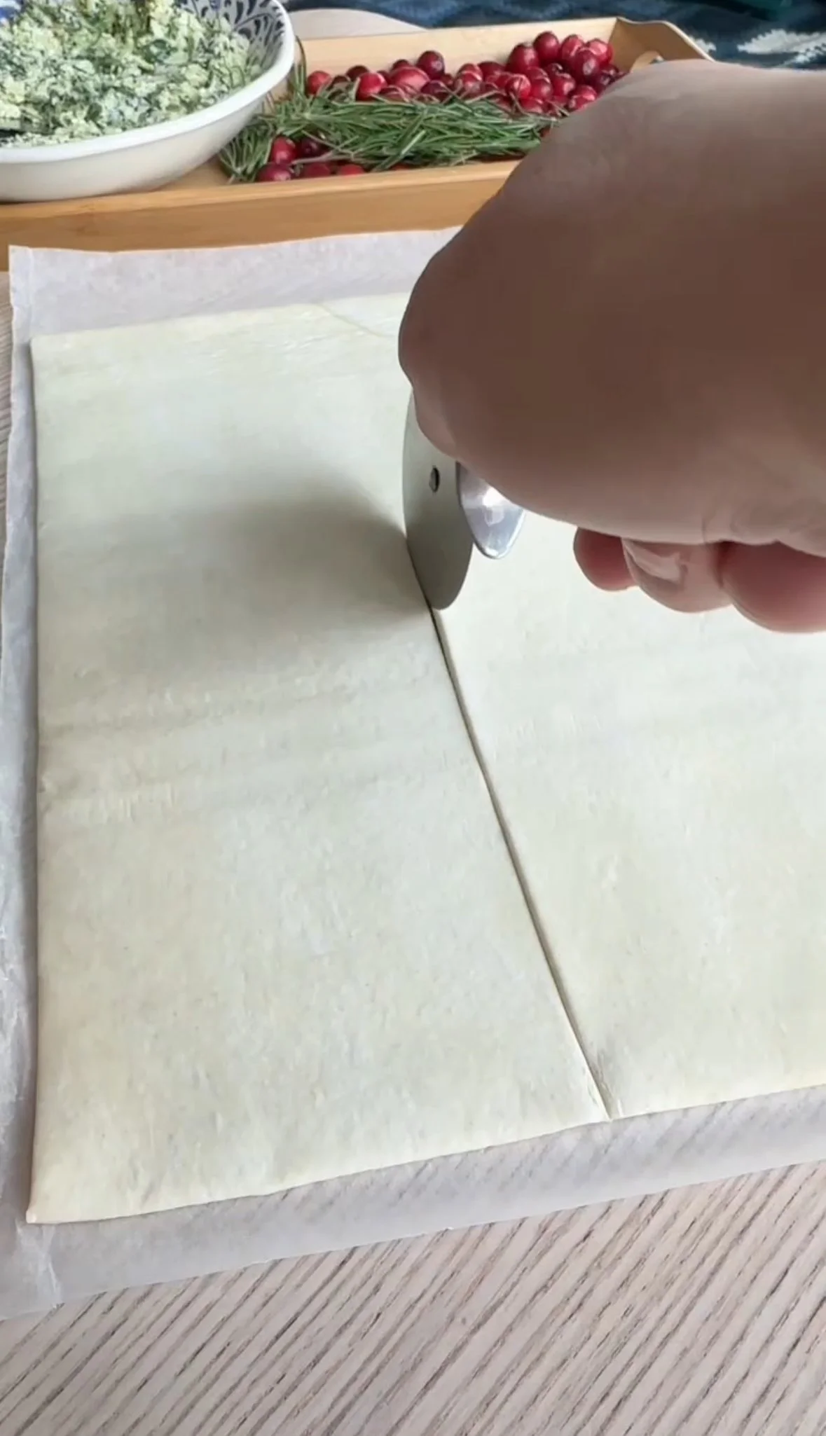 Person cutting a sheet of puff pastry with a round metal cutter, with fresh ingredients like cherries, radishes, and herbs visible in the background.