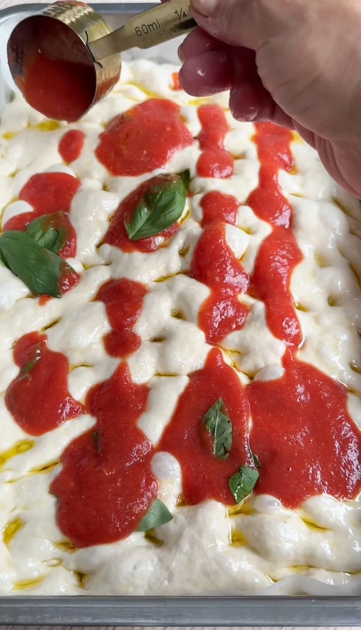 Close-up of a baking dish with layered mozzarella cheese, tomato sauce, and fresh basil leaves, with a hand drizzling more tomato sauce on top using a measuring cup.
