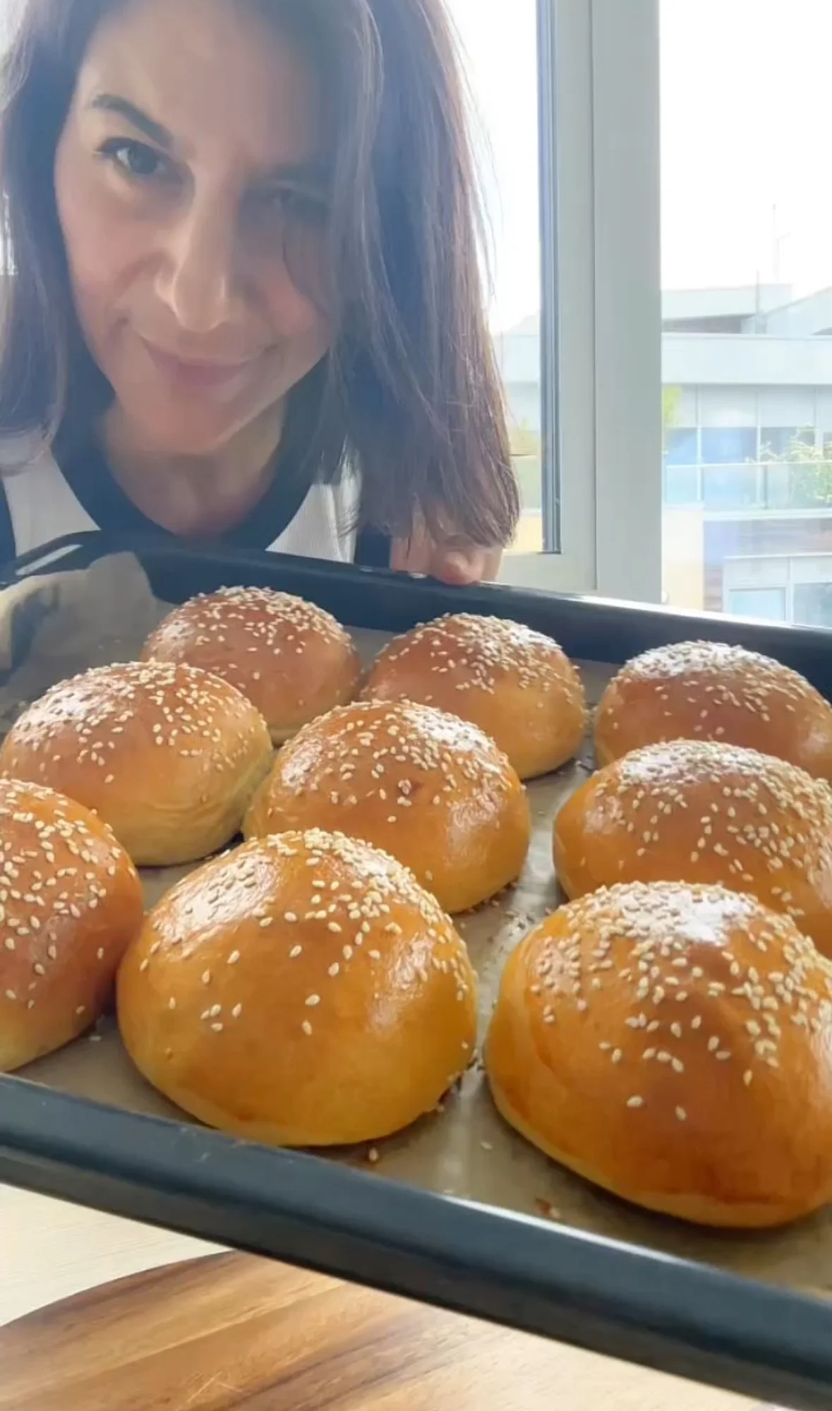 Woman smiling behind a tray of freshly baked sesame seed buns near a window.
