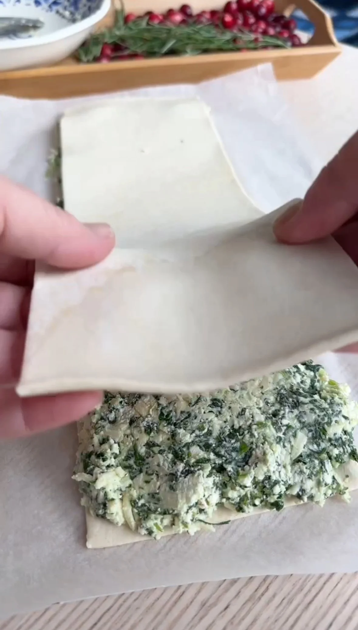 Someone is folding a square piece of pastry over a spinach and cheese filling on a sheet of parchment paper, preparing a pastry dish.