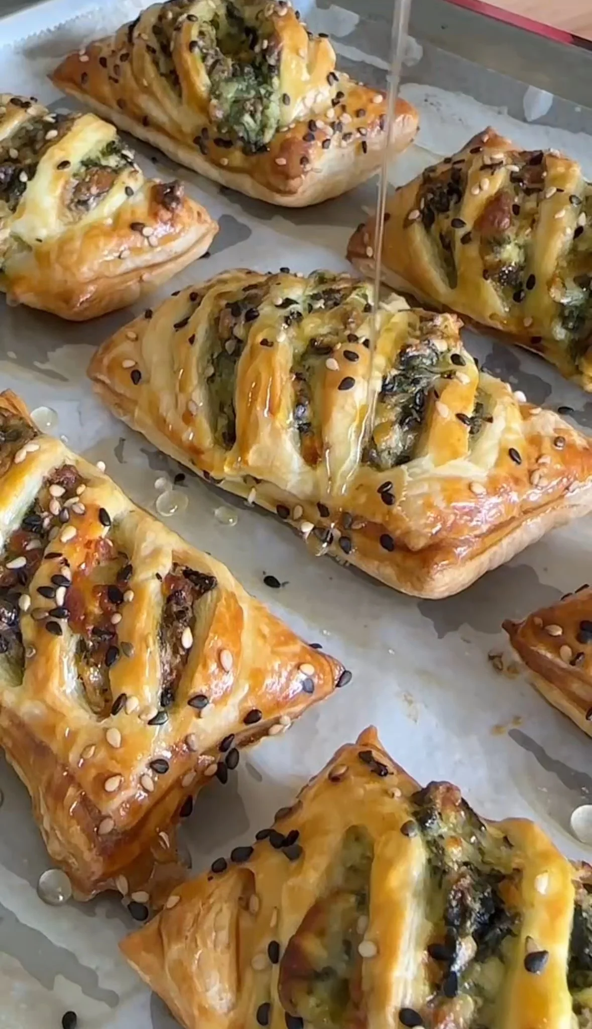 Close-up of savory baked pastries with a golden crust, topped with sesame and black seeds, filled with green vegetables and cheese, with a drizzle of honey.
