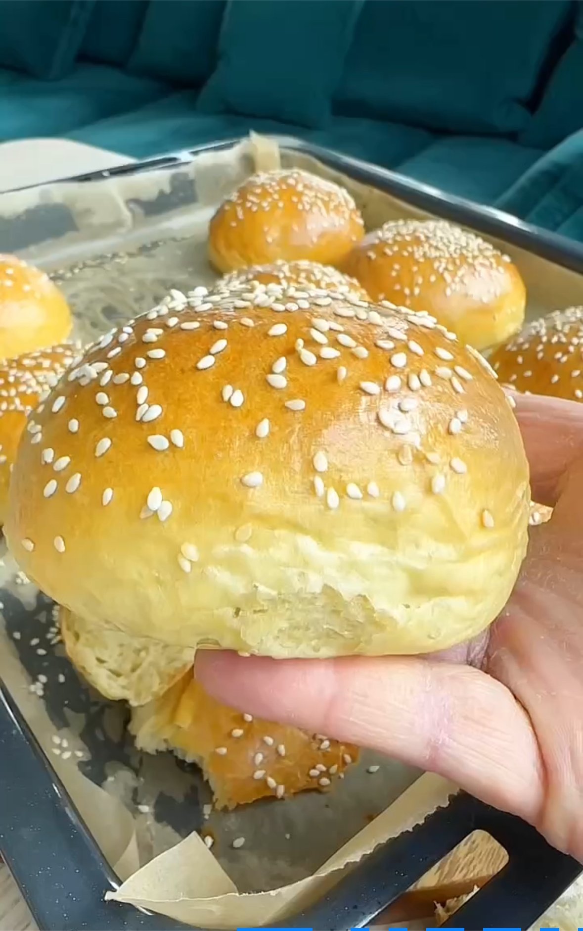 Close-up of a person's hand holding a freshly baked, sesame seed topped bread roll, with more similar rolls on a baking tray in the background.