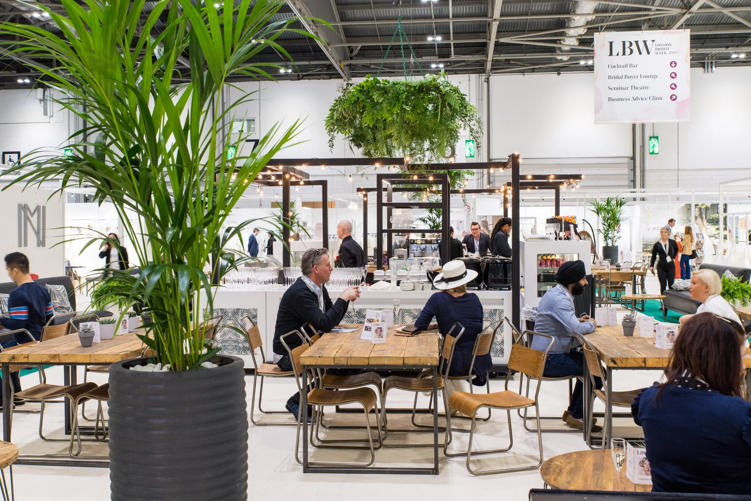 Indoor lounge live event space with tables, chairs, and people dining, large green potted plant in foreground, hanging greenery, and a bar area in the background. People networking