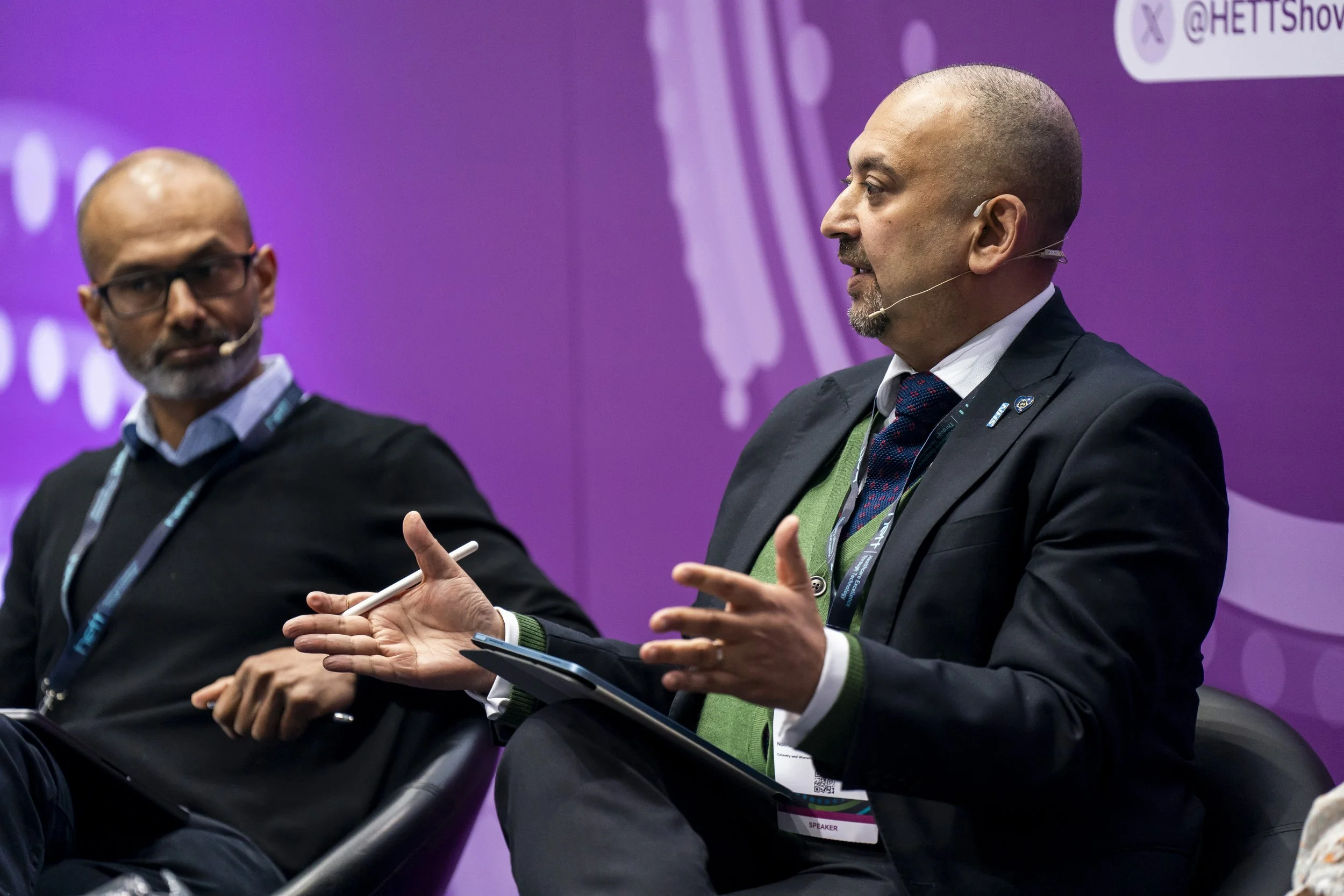 Two men engaging in a discussion on a panel at a conference, with a purple backdrop and company logos.