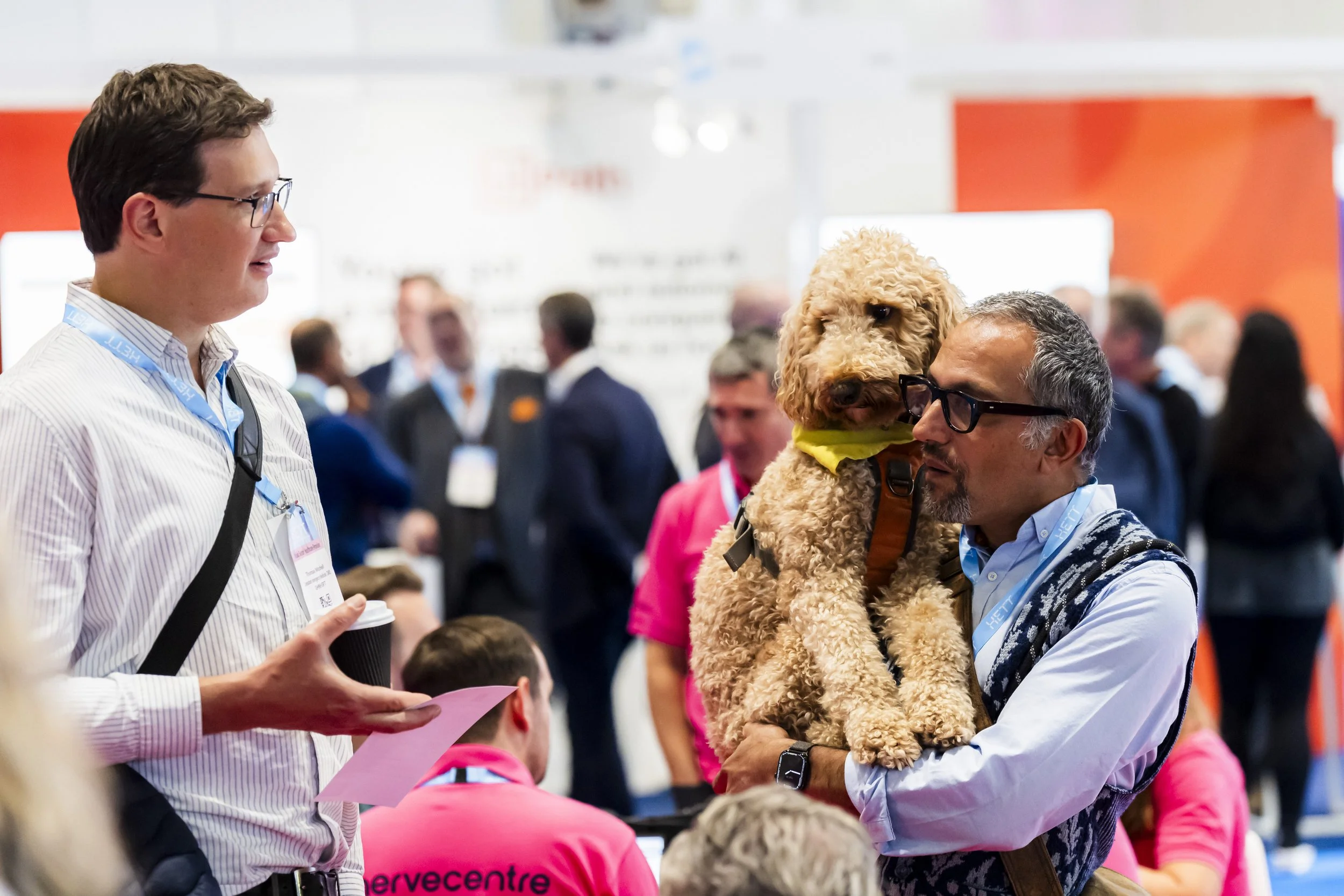 A man holding a curly-haired dog and a woman talking to him at a busy exhibition in the UK
