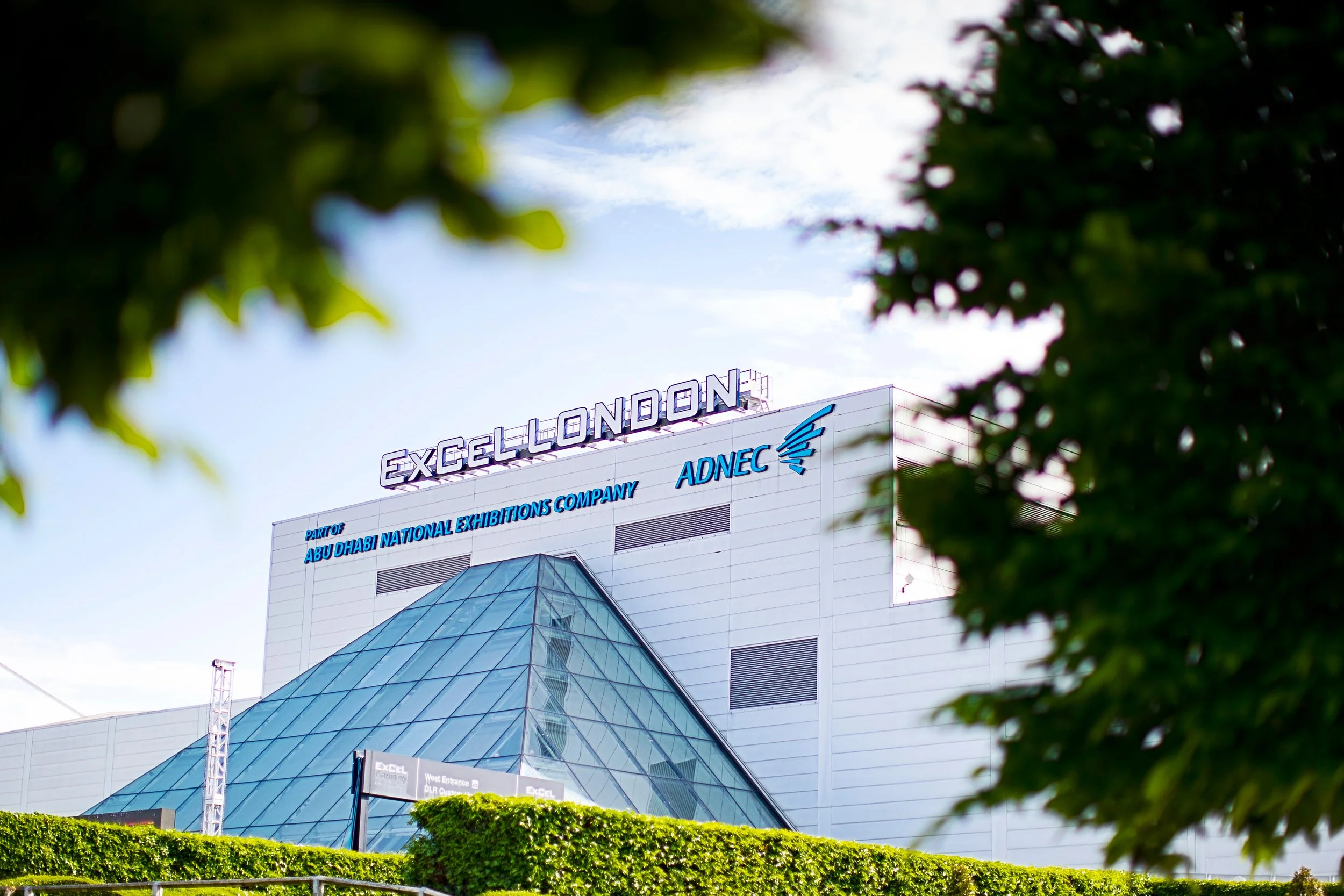 A modern venue with the sign 'EXCEL LONDON' on top, surrounded by green trees and bushes, under a partly cloudy sky. Excel London is an Adnec owned venue