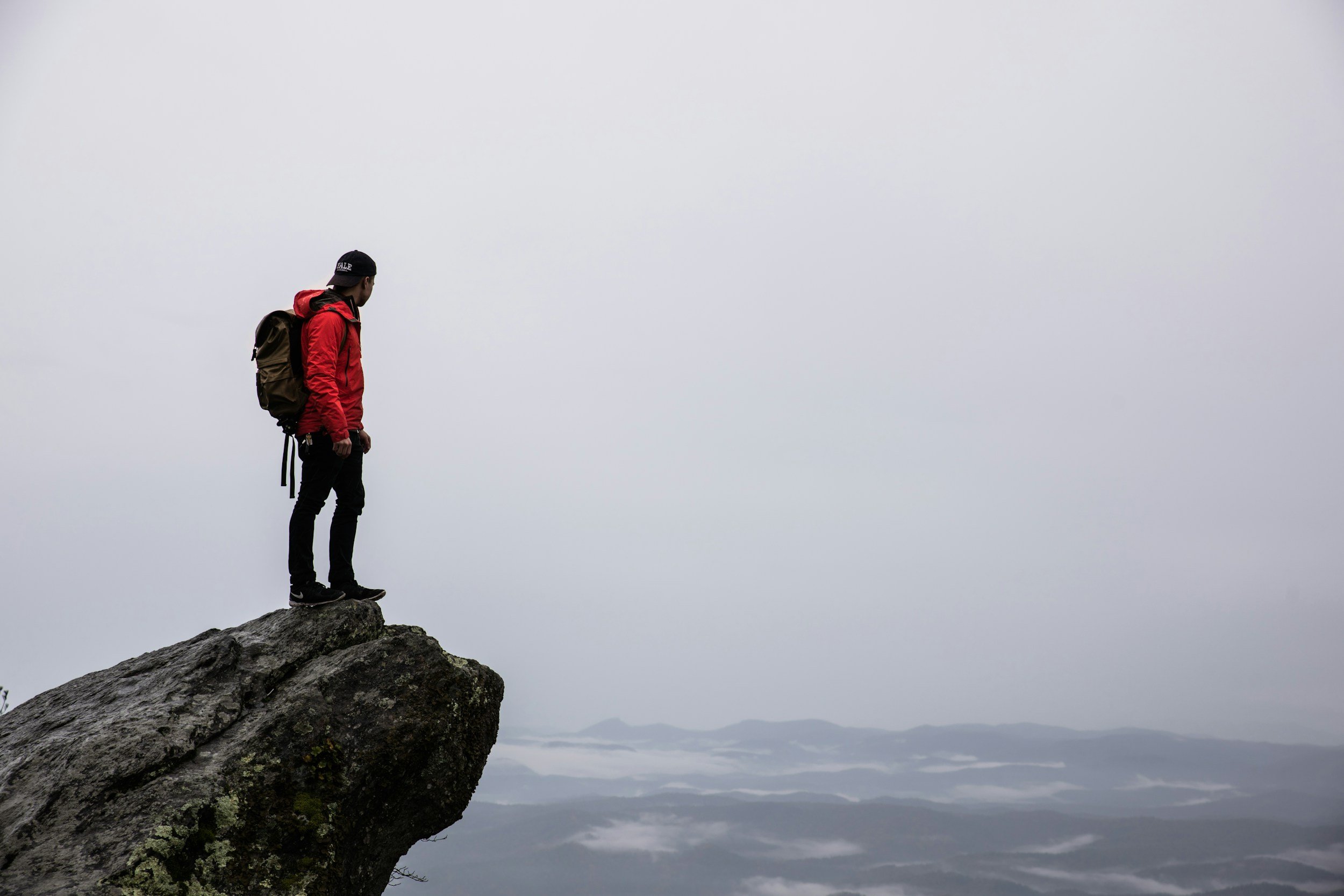 A person wearing a red jacket, black pants, and a black cap with a backpack, standing on the edge of a rocky cliff overlooking a foggy landscape with mountains and clouds.
